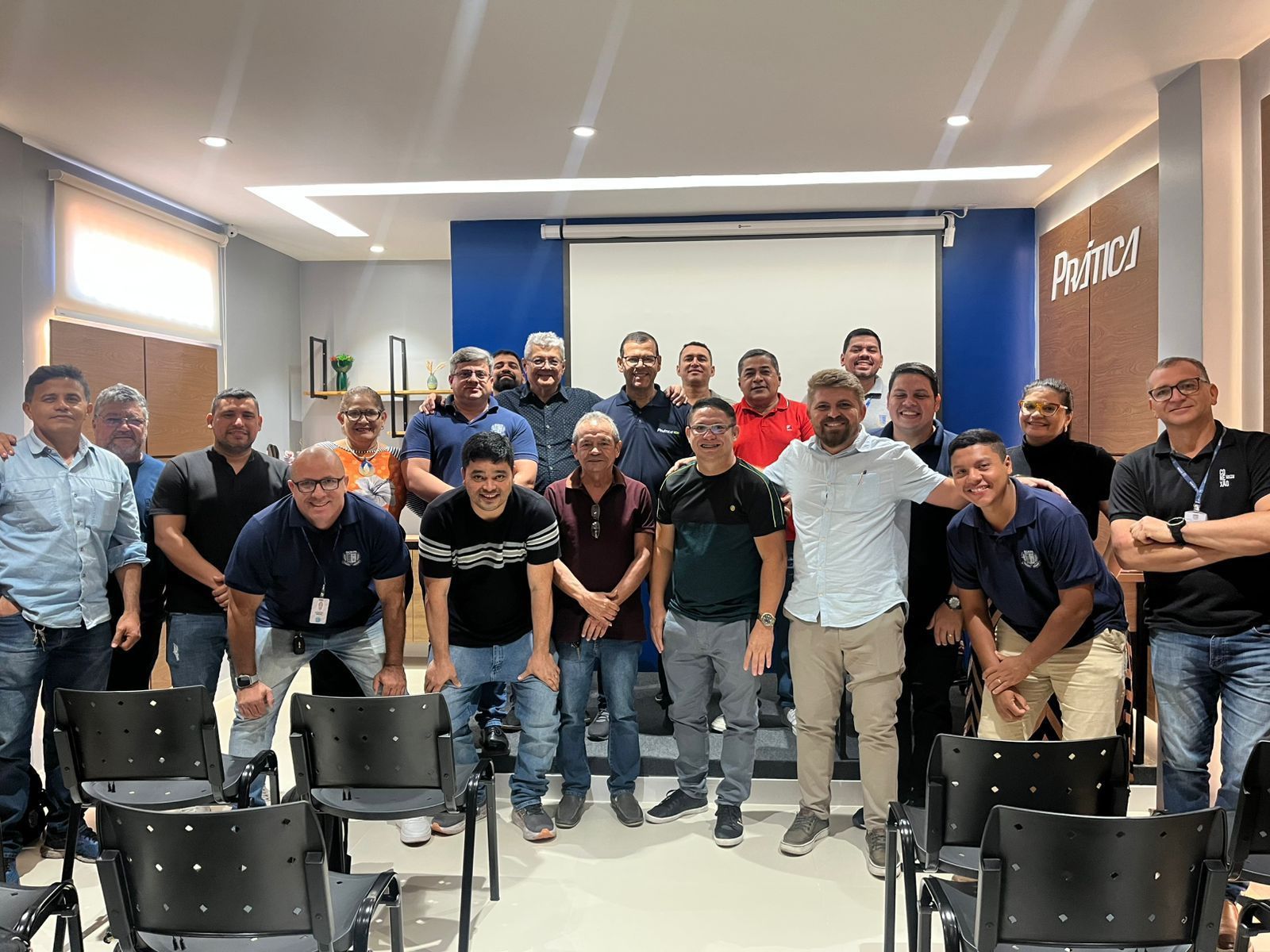 Grupo de homens em uma sala de reuniões, sorrindo, posando para uma foto; cenário azul, cinza e branco.