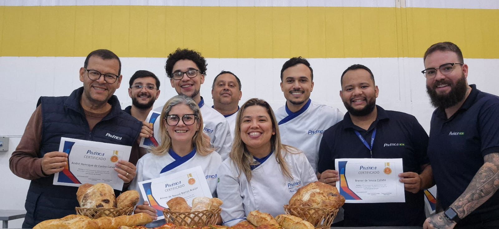 Um grupo de pessoas, algumas segurando certificados, sorriem atrás de uma mesa de produtos assados em uma cozinha.