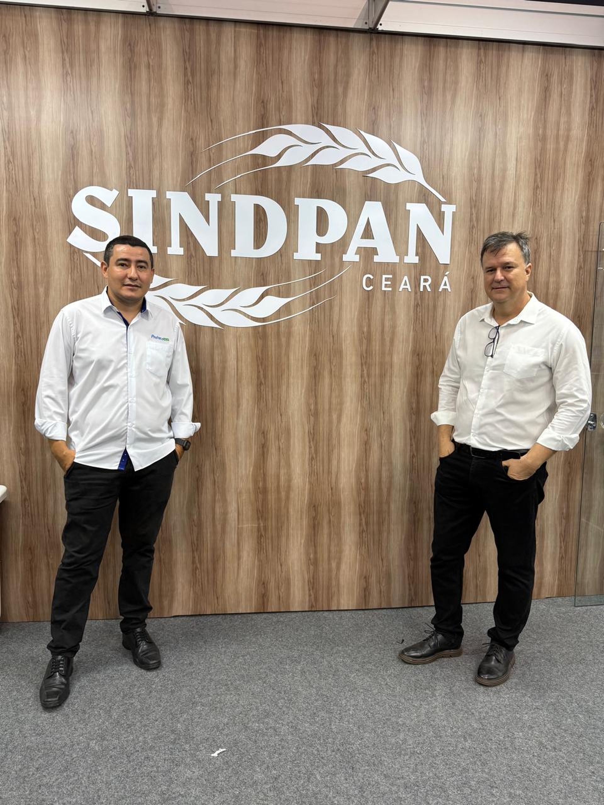 Duas pessoas estão lado a lado em frente a uma parede de madeira com o logotipo 