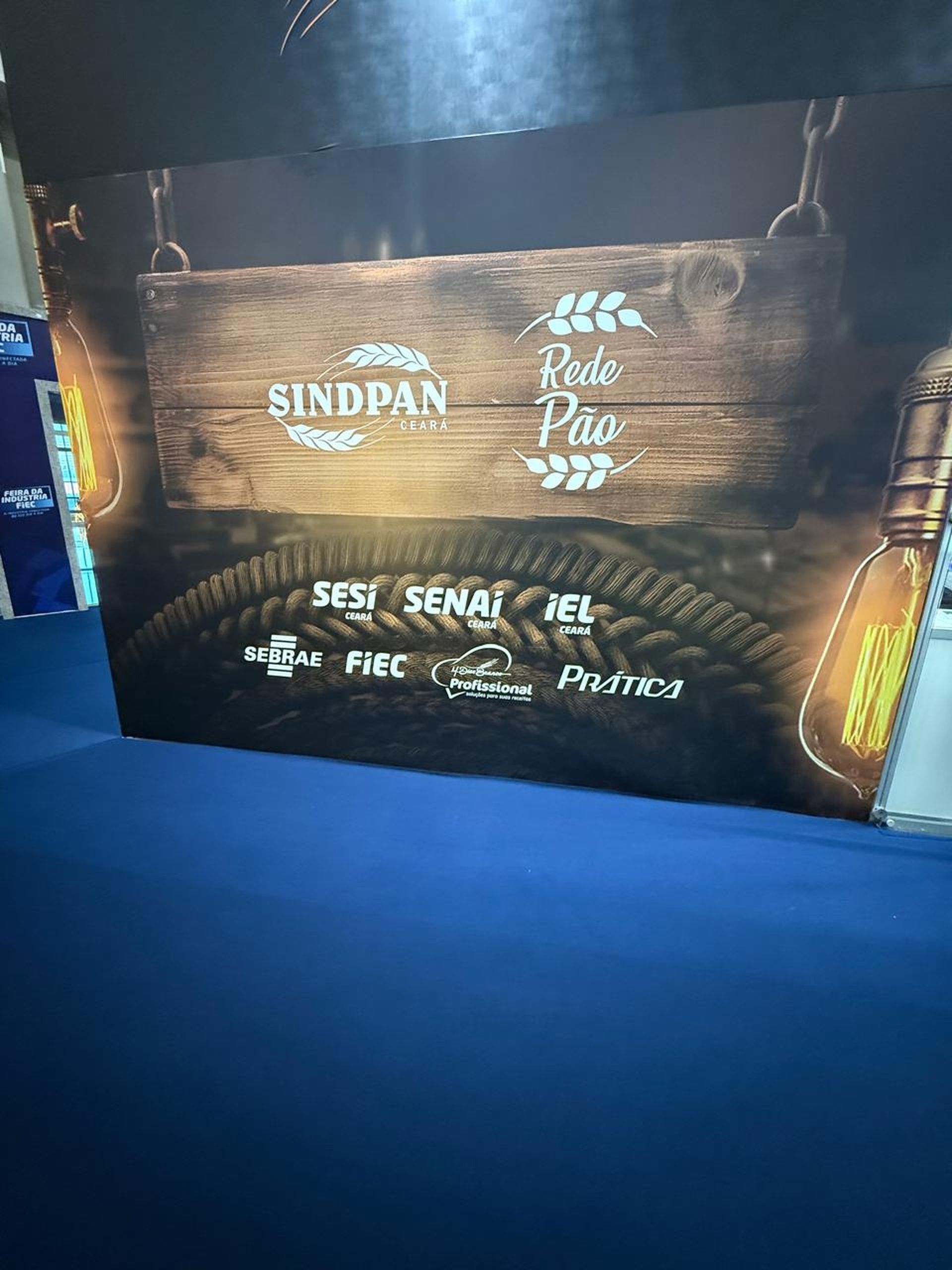 Uma placa de madeira pendurada por correntes contra um fundo escuro, exibindo os logotipos da Sindpan