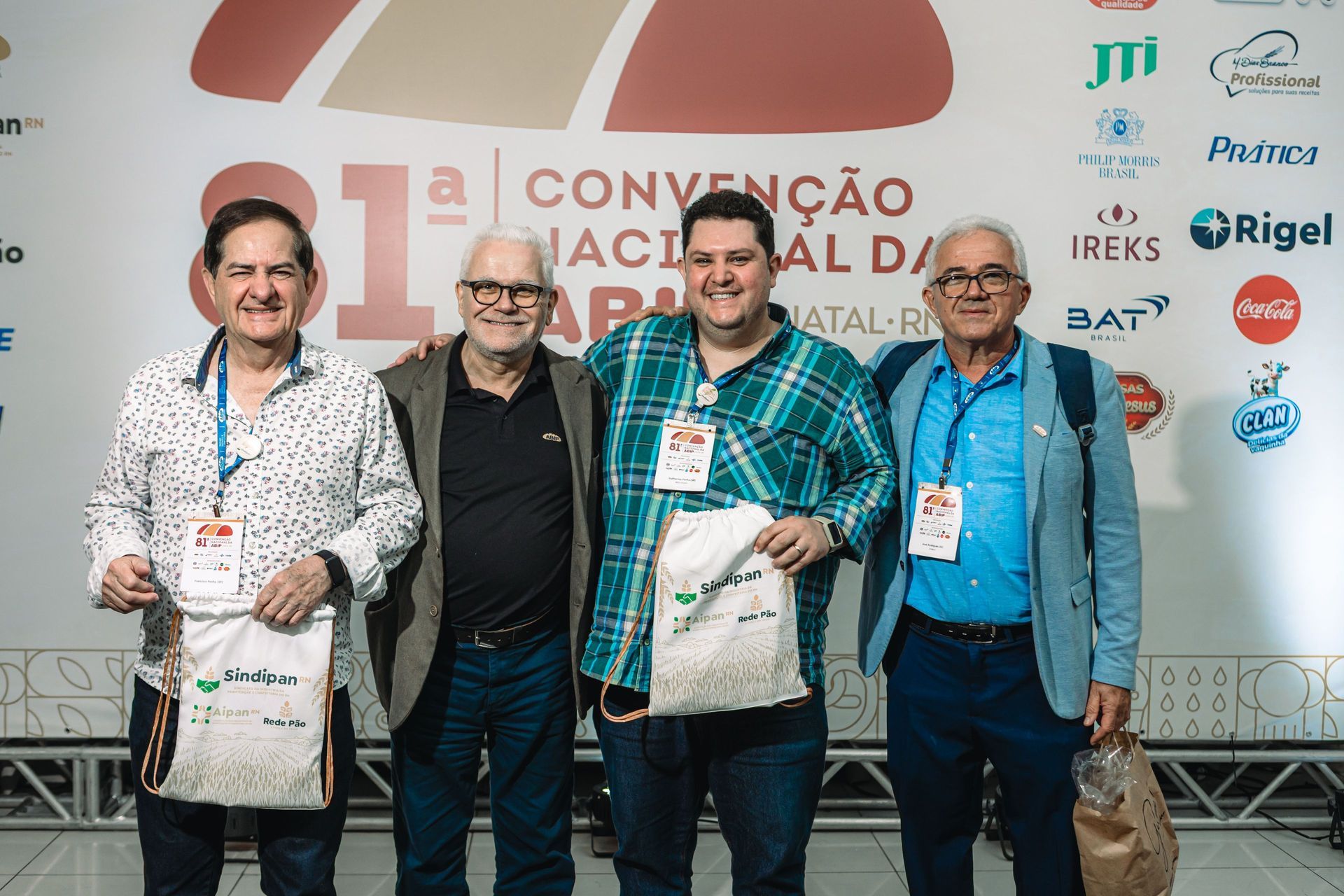 Quatro homens posam para foto em uma convenção, cada um segurando uma bolsa e usando um crachá.