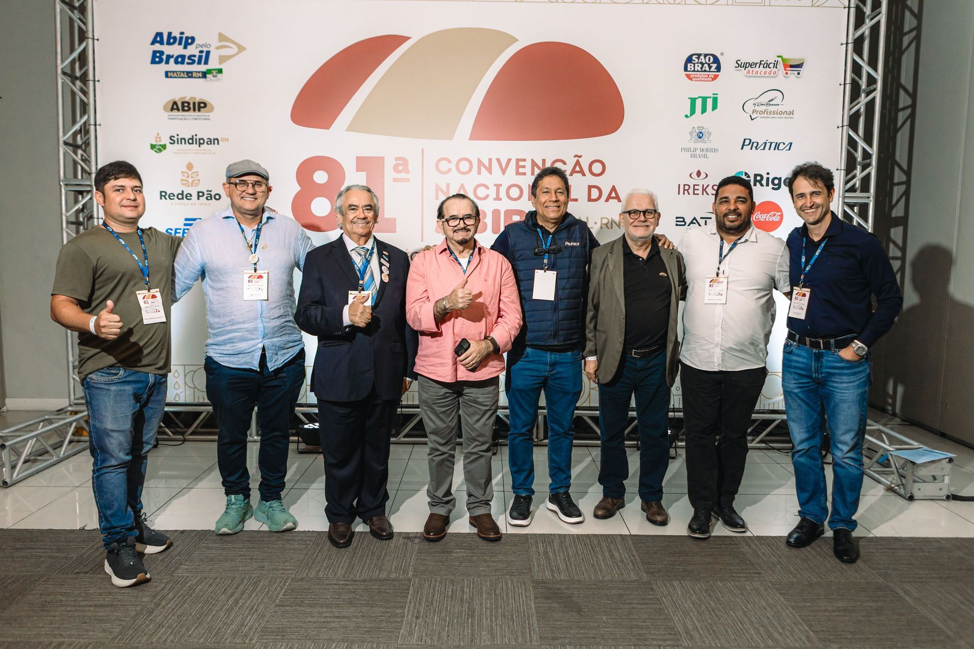 Grupo de homens em uma convenção. Eles estão em frente a uma faixa com texto e logotipos, sorrindo e posando para uma foto.