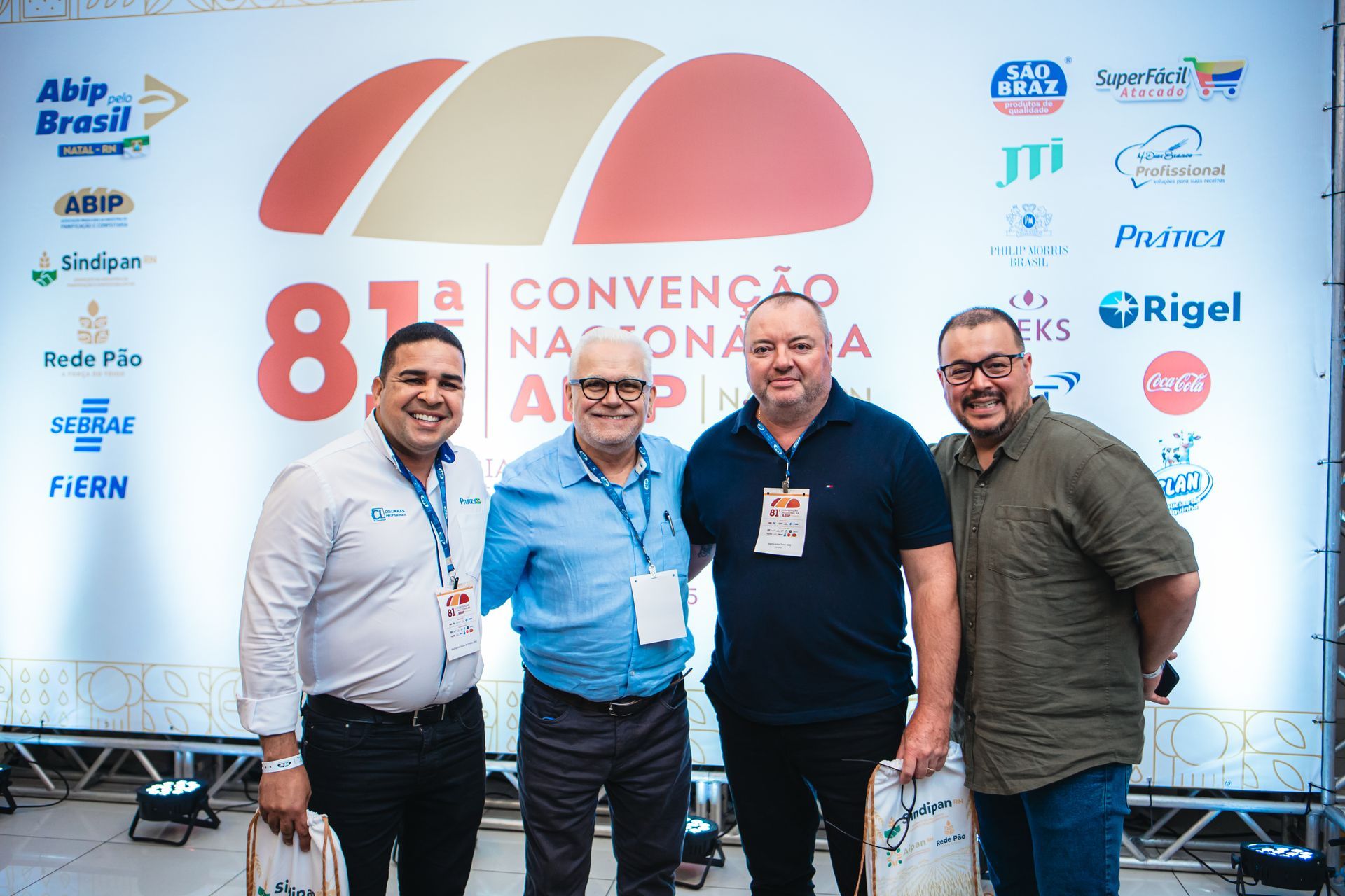 Quatro homens posam em frente a um painel de fundo de conferência com logotipos e texto.