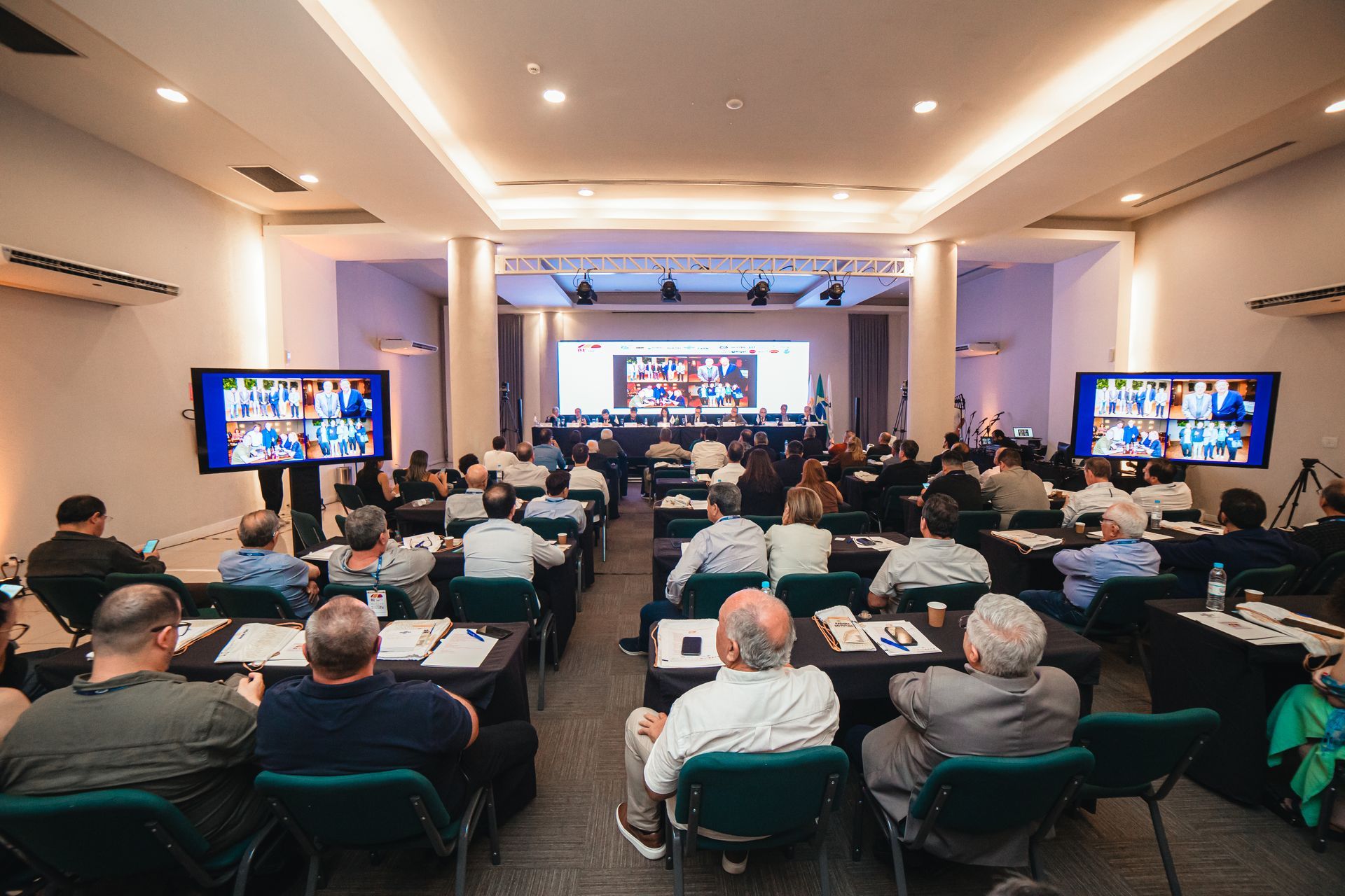 Sala de conferências com participantes, telas exibindo videochamadas e palco na frente.