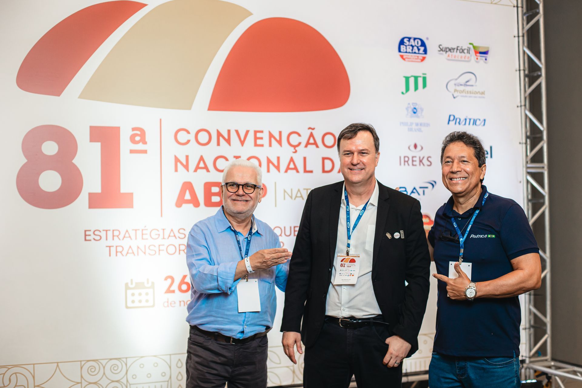 Três homens sorrindo em frente a um painel de fundo de convenção, possivelmente brasileiro. Um deles aponta para a faixa.
