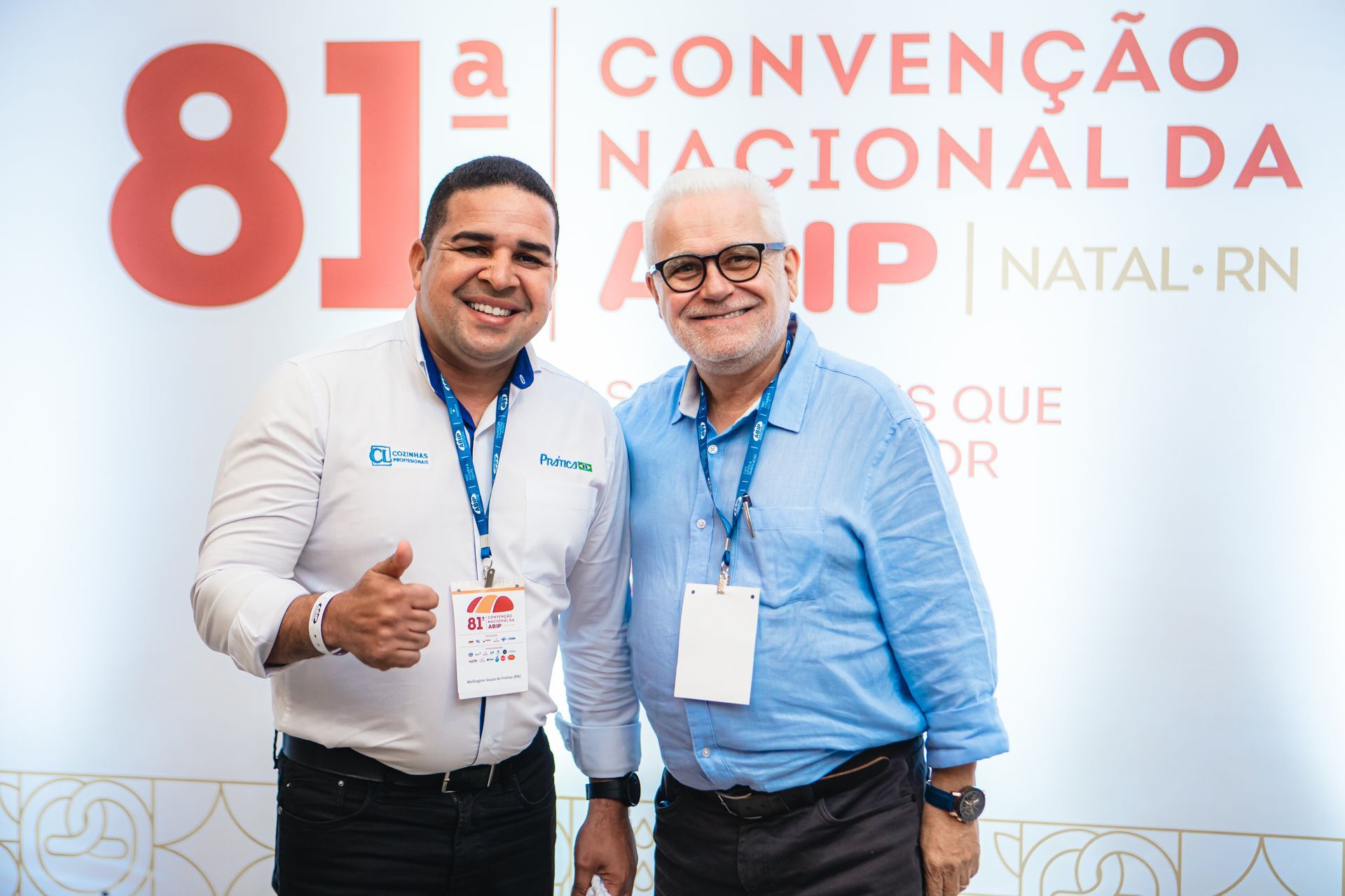 Dois homens sorrindo, posando para uma foto em uma conferência em Natal, RN. Um deles faz um sinal de positivo com o polegar. Uma faixa diz 