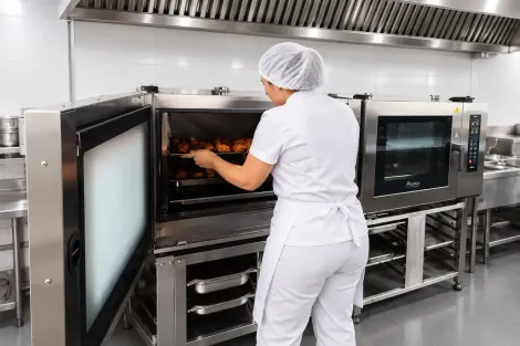 Trabalhador colocando bandejas em um grande forno industrial de aço inoxidável em uma cozinha comercial