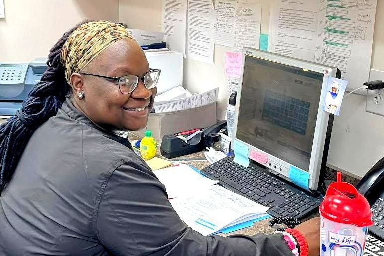 Woman at computer smiling, in an office setting. Wearing glasses and a headscarf, looking towards the camera in Eastover, NC.