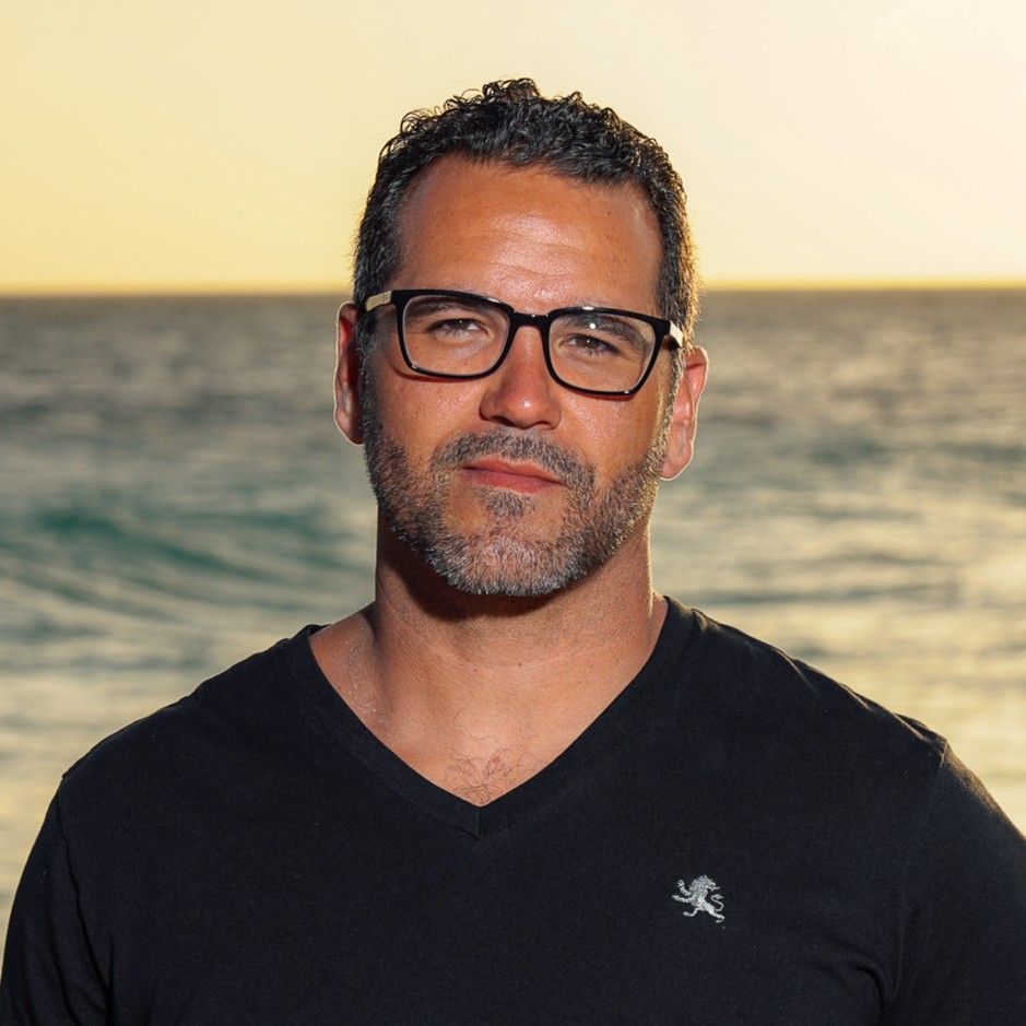 Man wearing glasses and a black V-neck shirt standing by the ocean at sunset