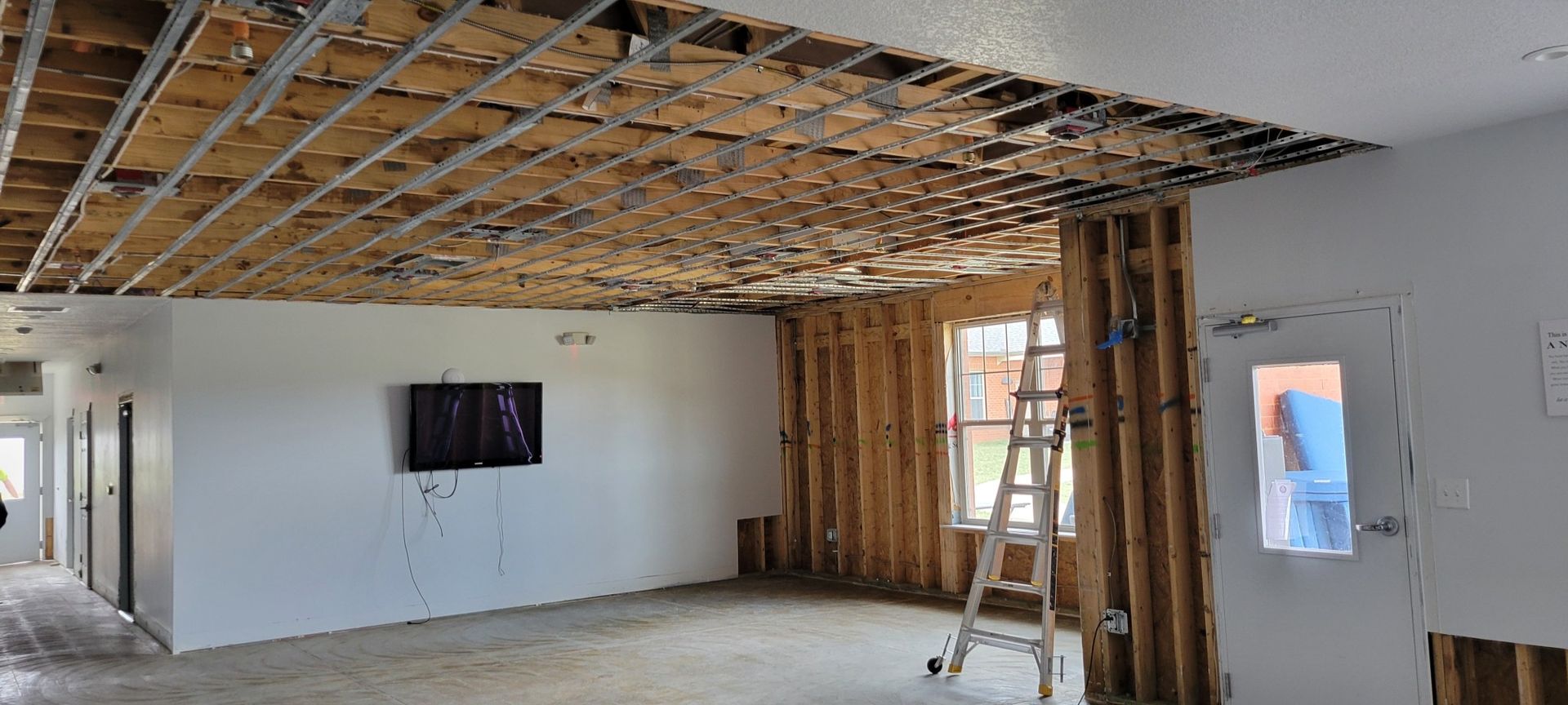 Interior view of a damaged room. Ceiling is partially collapsed; walls are exposed studs. A ladder stands near a window.