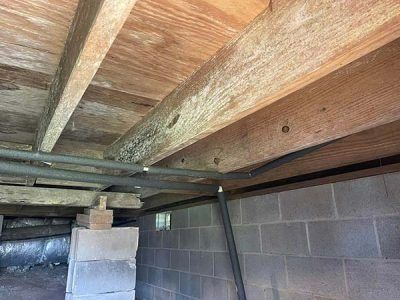 Underneath a house: wooden beams, grey pipes, cinder block foundation, and supports.
