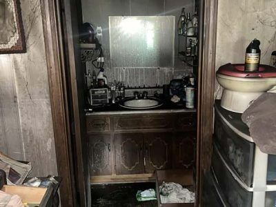 Dirty, cluttered bathroom with a sink, toilet, and cabinet. Brown walls and fixtures are visible.
