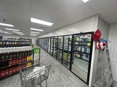 Interior of a convenience store with refrigerated glass doors, shelving, and a shopping cart.