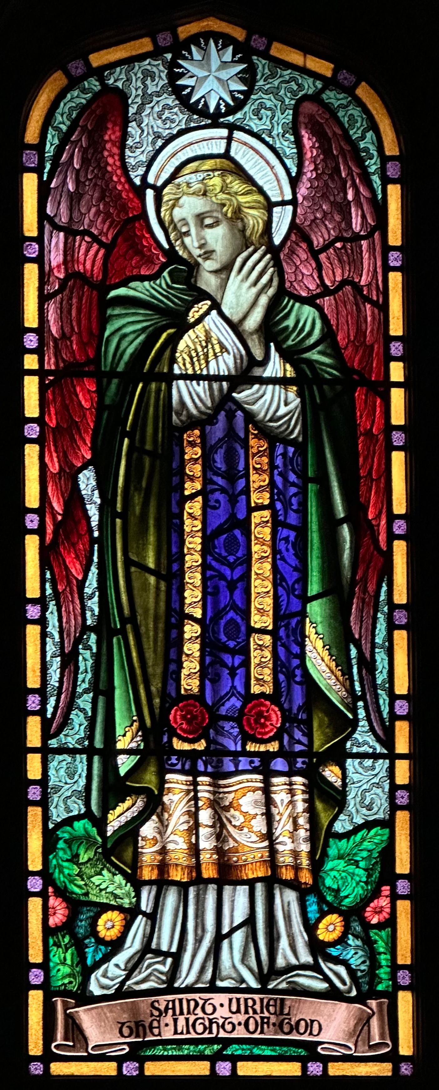 Stained glass angel with red wings, praying hands, and a halo. Green and purple robes.