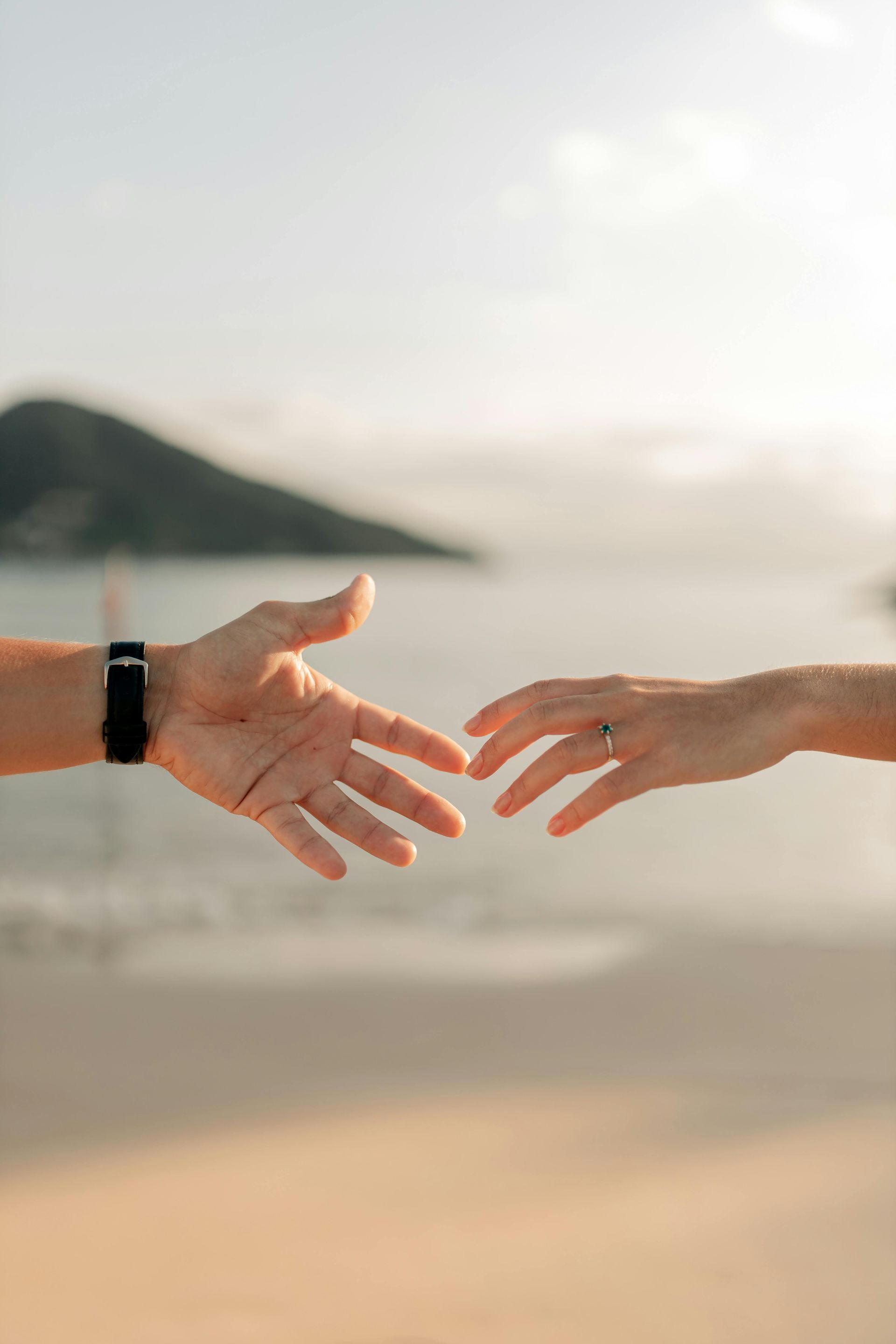 Image of two hands reaching out towards each other in the foreground of a beach scene. 