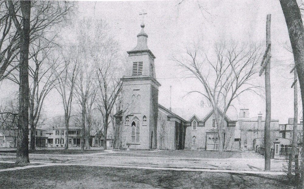 Black and white photo of a tall church with a steeple, trees in front, and buildings in the background.