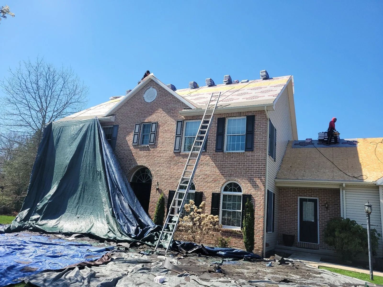 House roof repair with ladders, tarps, and workers on the roof under a clear blue sky
