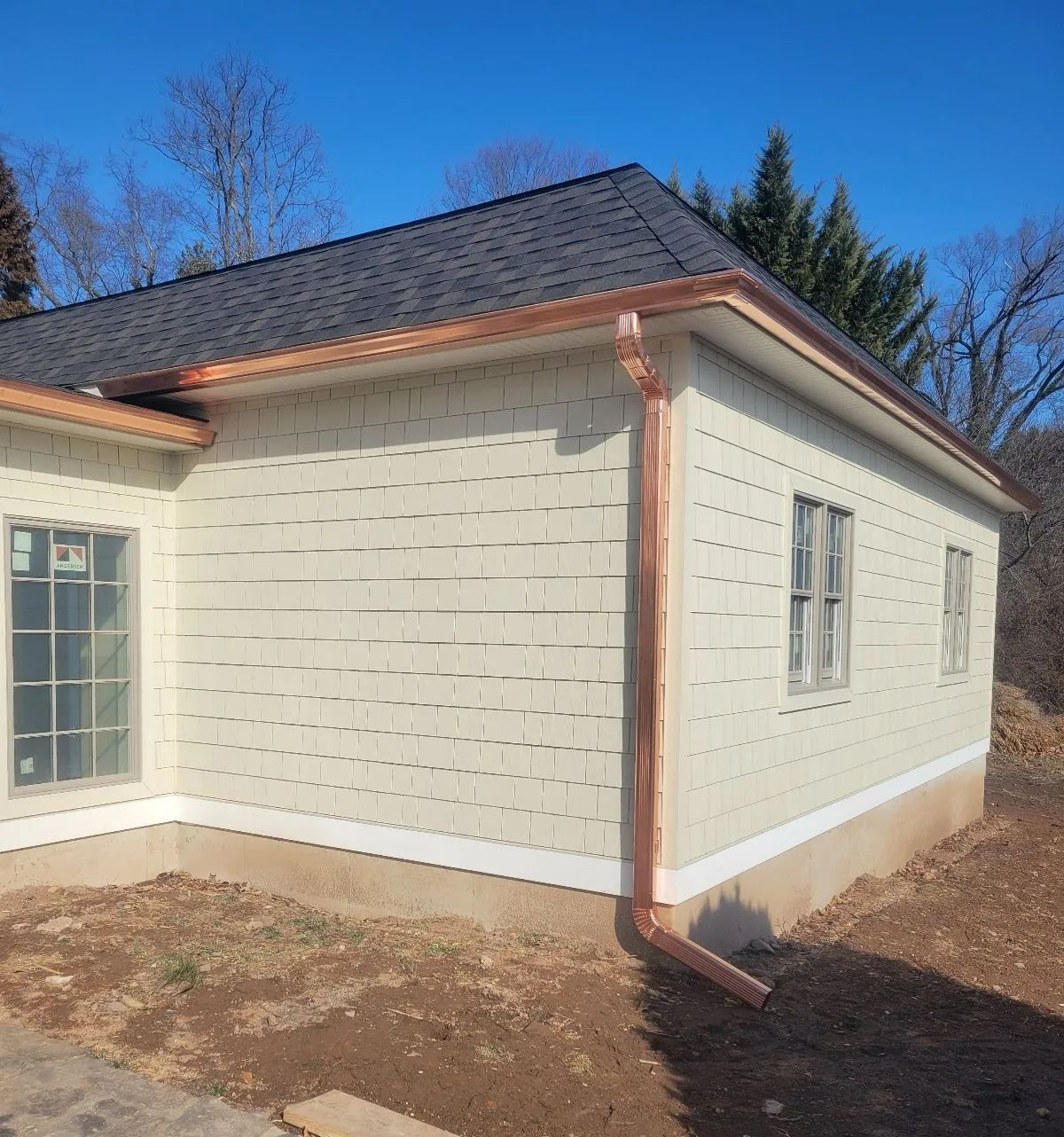 Exterior corner of a beige house with white trim, dark shingled roof, and copper downspout