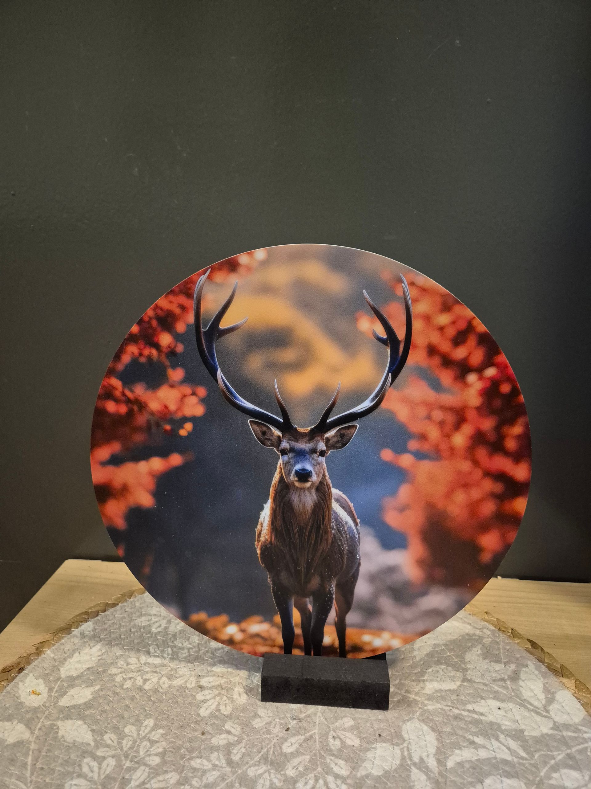 Rond kunstwerk van een hert met gewei tegen een warme herfstbosachtergrond, gepresenteerd op een displaystandaard.