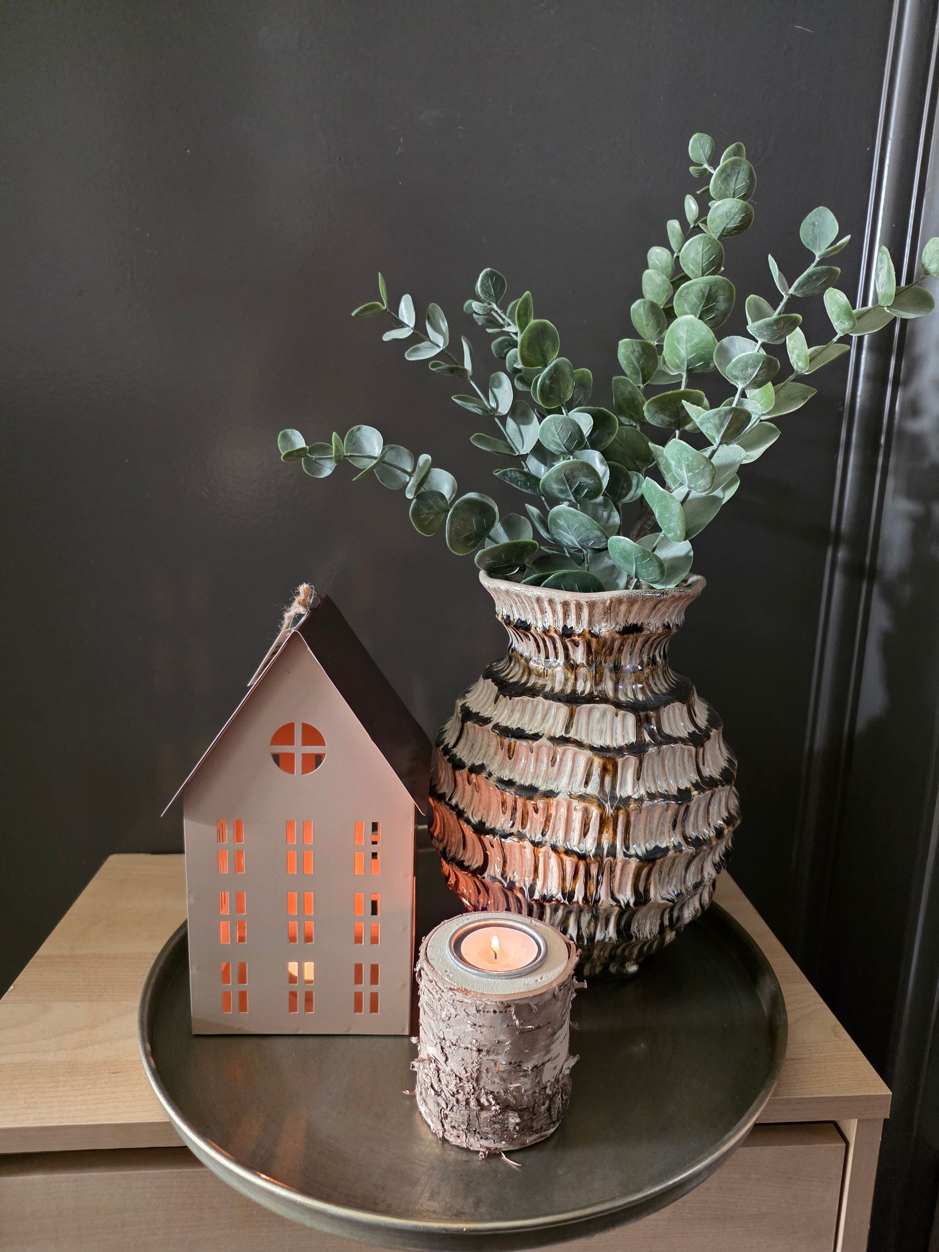 Rustiek kaarsenarrangement met een kleine lantaarn in de vorm van een huisje en eucalyptus in een vaas met textuur op een dienblad.
