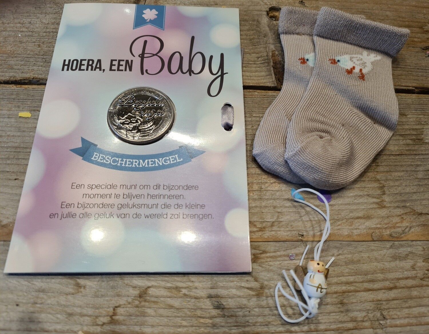 Babycadeauset met een kaartje, zilveren speld, grijze babysok en een klein hangend ornamentje op een houten ondergrond.