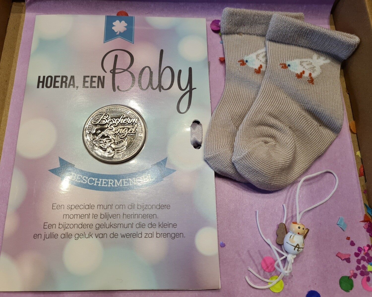 Babycadeauset met een kaartje, zilveren speld, grijze babysok en een klein hangend ornamentje op een houten ondergrond.