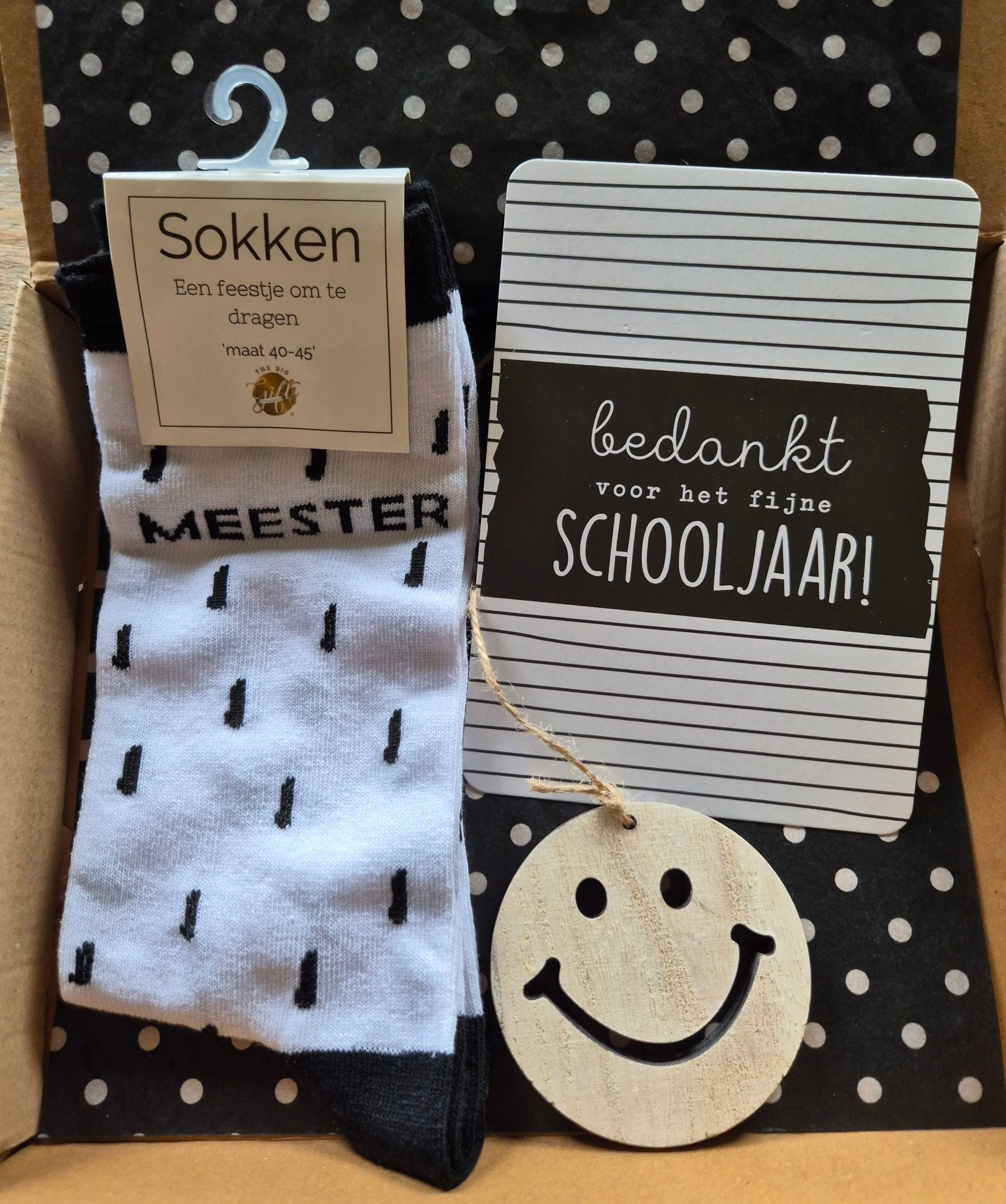 Gift set with socks, “meester” sign, “bedankt schooljaar!” card, and smiley ornament on a polka-dot background