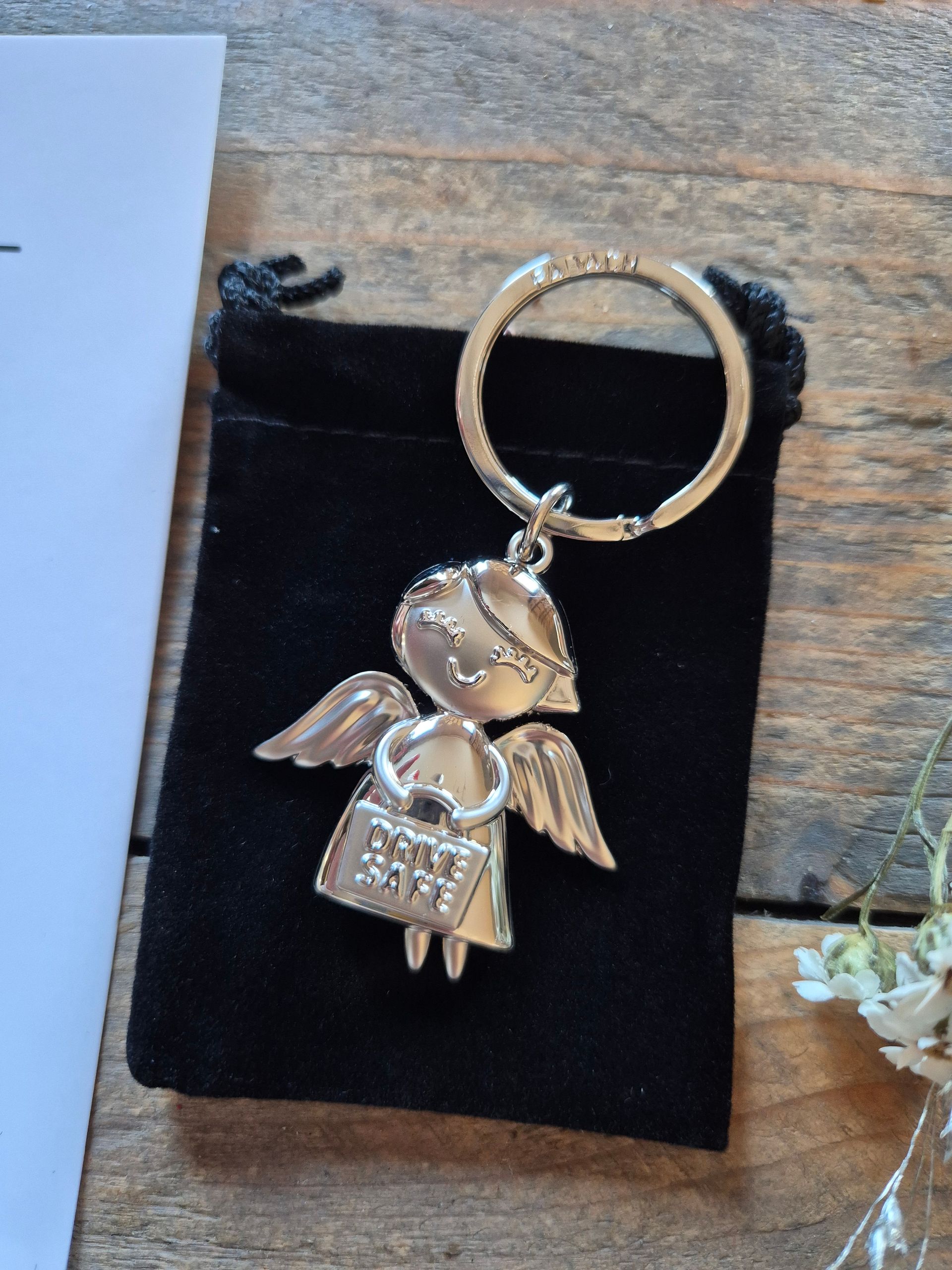 Cadeaukaart met auto-icoon en Nederlandse tekst naast een gouden engelsleutelhanger op een zwarte kaart, naast gedroogde bloemen.