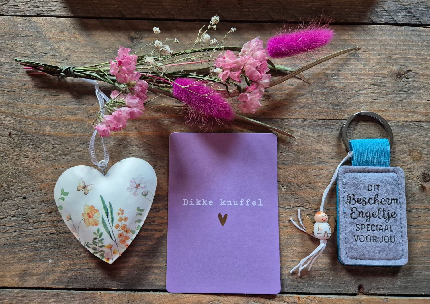 Cadeauset met roze bloemen, een hartvormig ornament, een lavendelkleurig kaartje en een kleine, gepersonaliseerde flesopener op een stoffen doek met stippen.