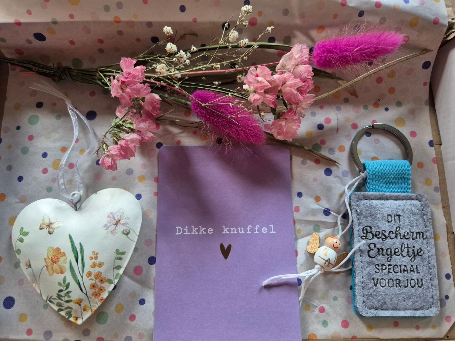 Cadeauset met roze bloemen, een hartvormig ornament, een lavendelkleurig kaartje en een kleine, gepersonaliseerde flesopener op een stoffen doek met stippen.