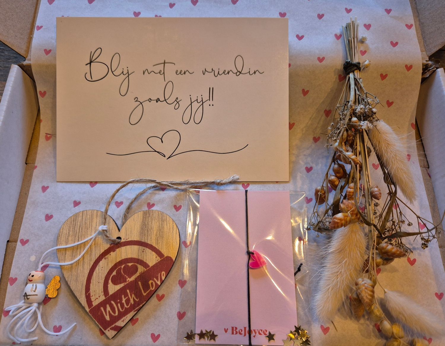 Cadeaupresentatie met handgeschreven briefje, hartvormige ornamenten en een klein versierd kerstboompje op een pastelkleurige achtergrond.