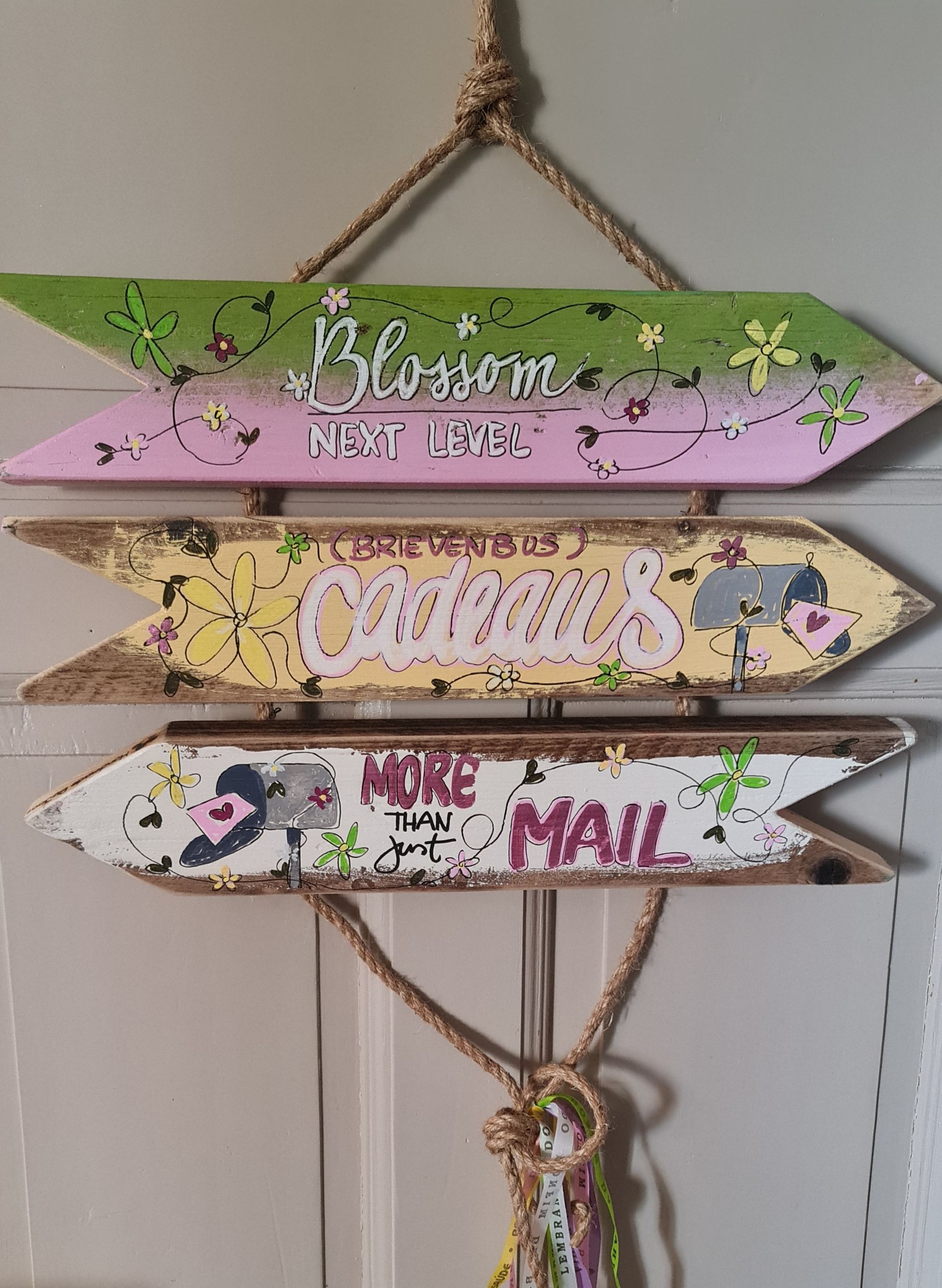 Drie pastelkleurige houten pijlvormige bordjes aan een touw, beschilderd met bloemen en woorden die de seizoenen en de post aangeven.