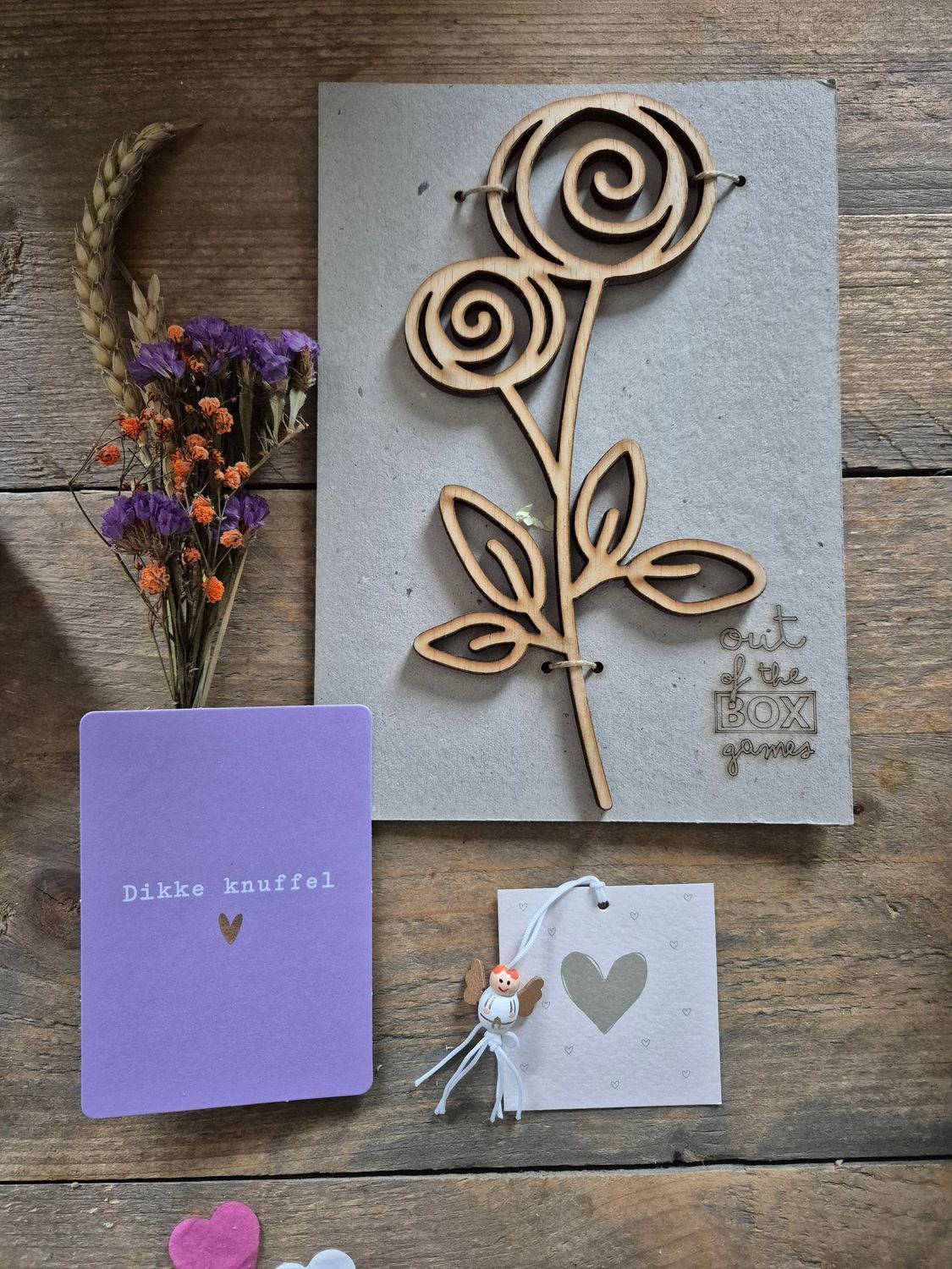 Cadeauverpakking met een spiraalvormige houten roos, een lavendelkleurig kaartje, gedroogde bloemen en een klein wit labeltje op een achtergrond met hartjespatroon.