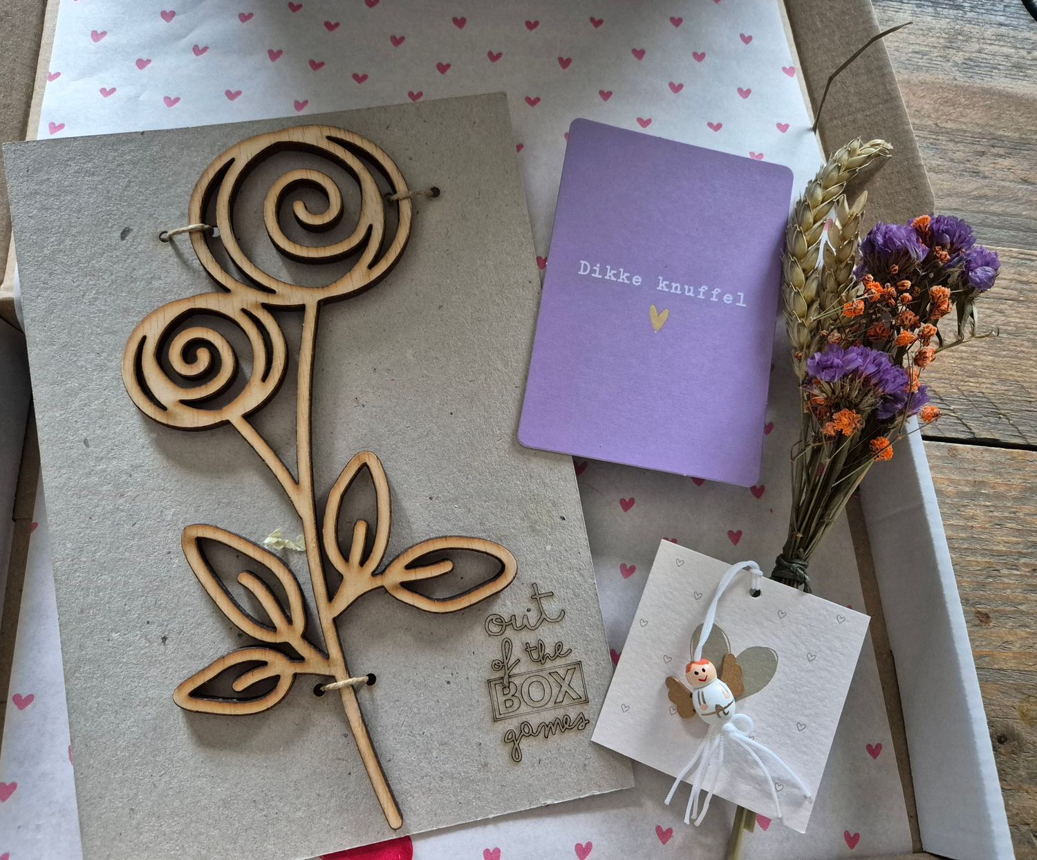 Cadeauverpakking met een spiraalvormige houten roos, een lavendelkleurig kaartje, gedroogde bloemen en een klein wit labeltje op een achtergrond met hartjespatroon.