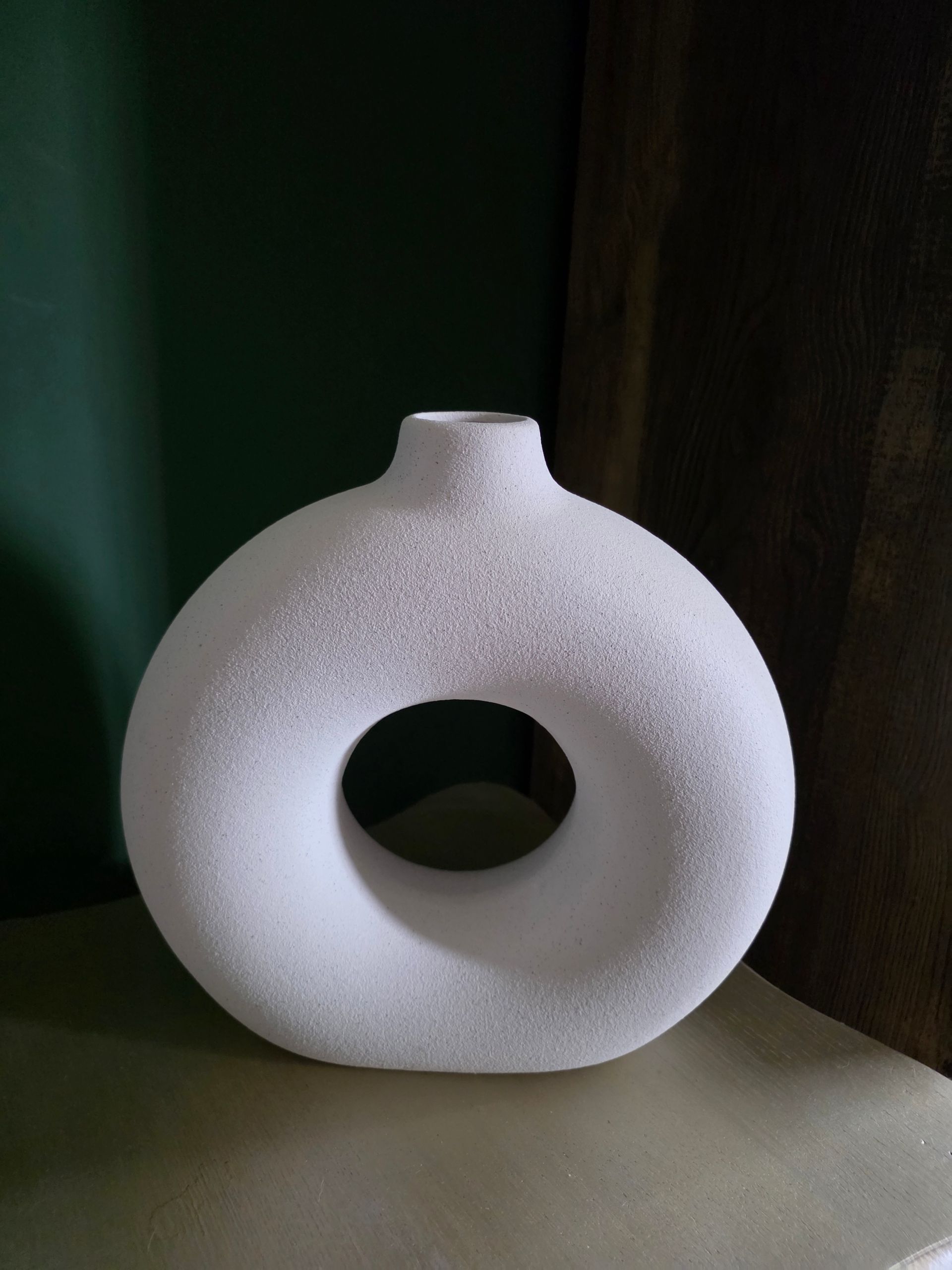 Witte keramische ringvormige vaas met een smalle hals op een plank, tegen een donkere achtergrond.