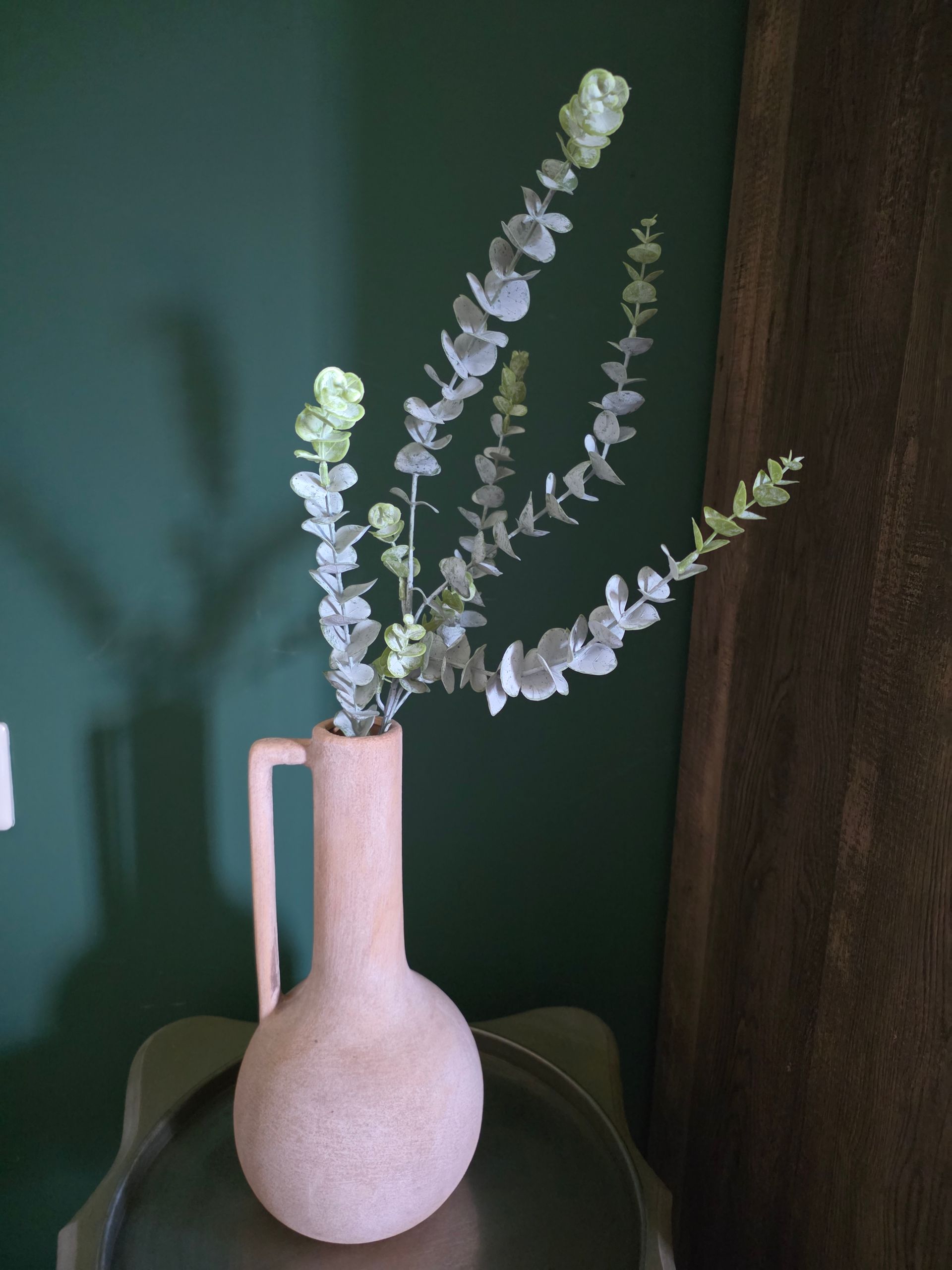 Witte vaas met eucalyptus takken op een tafel tegen een donkergroene muur.