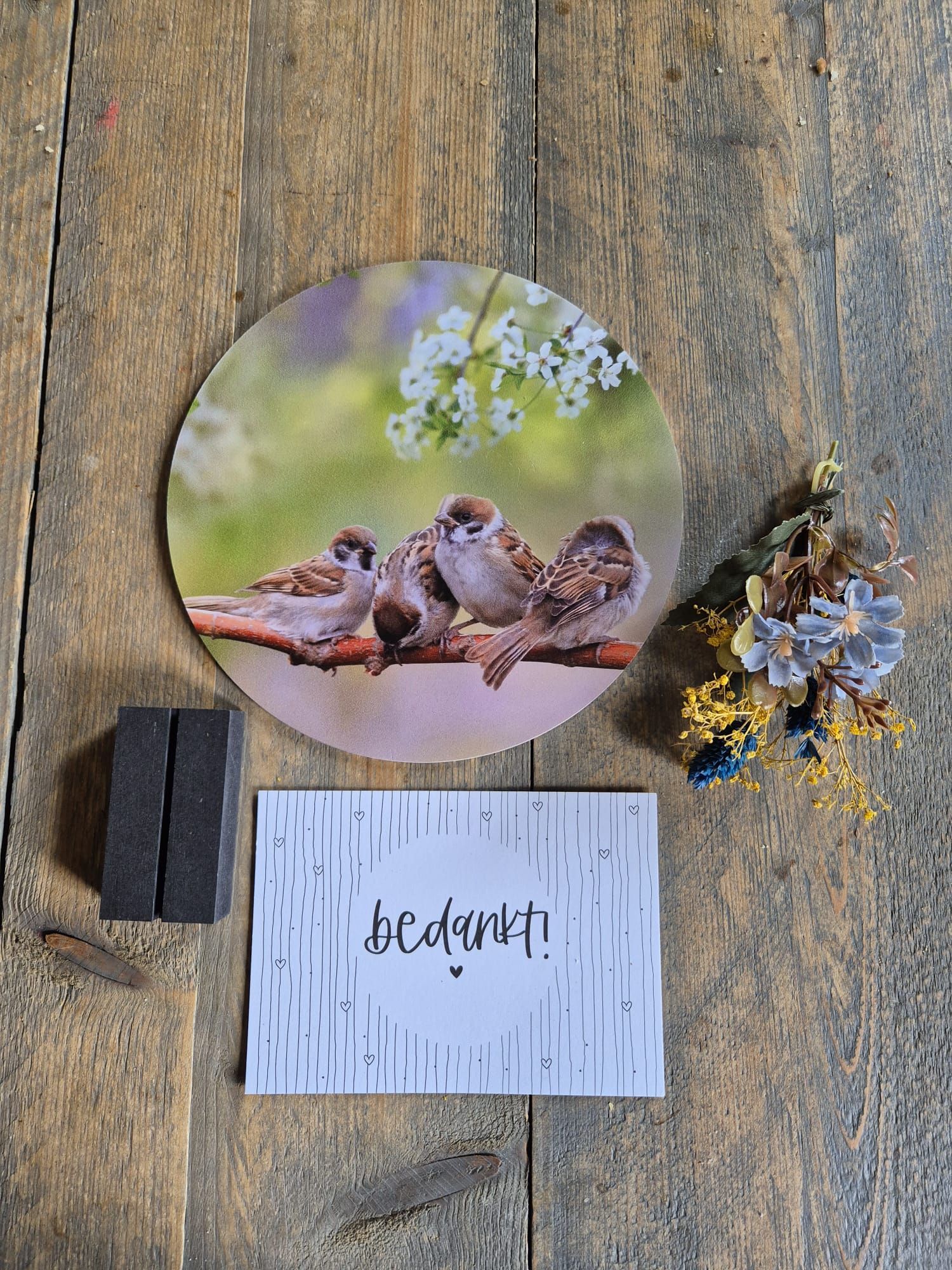 Rond fotobord met een afbeelding van twee mussen, een houten clip en gedroogde bloemen op een rustieke tafel.