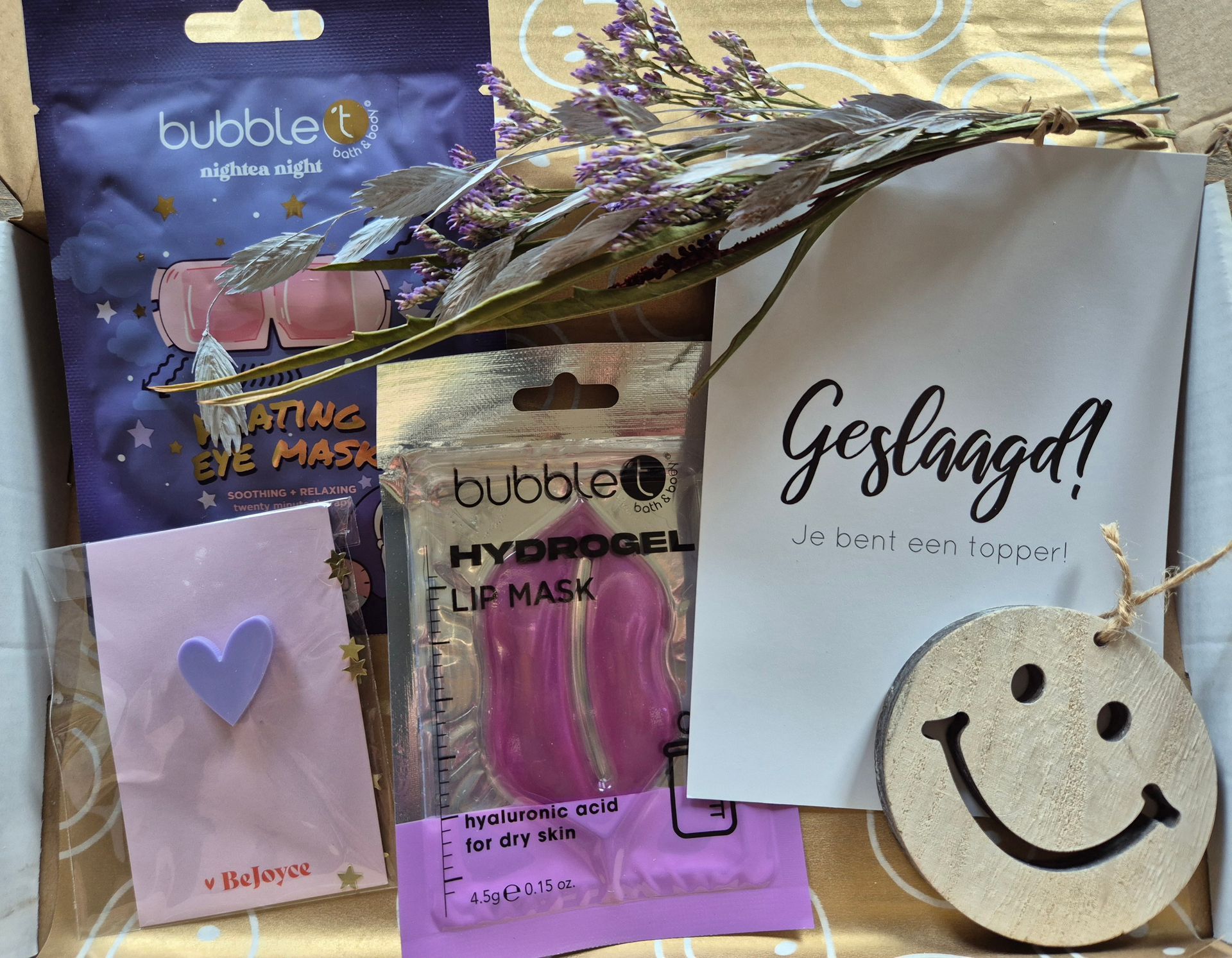 Diverse cosmetica- en cadeauartikelen op een bureau, waaronder een label met een smiley, een paars etui en gedroogde bloemen.
