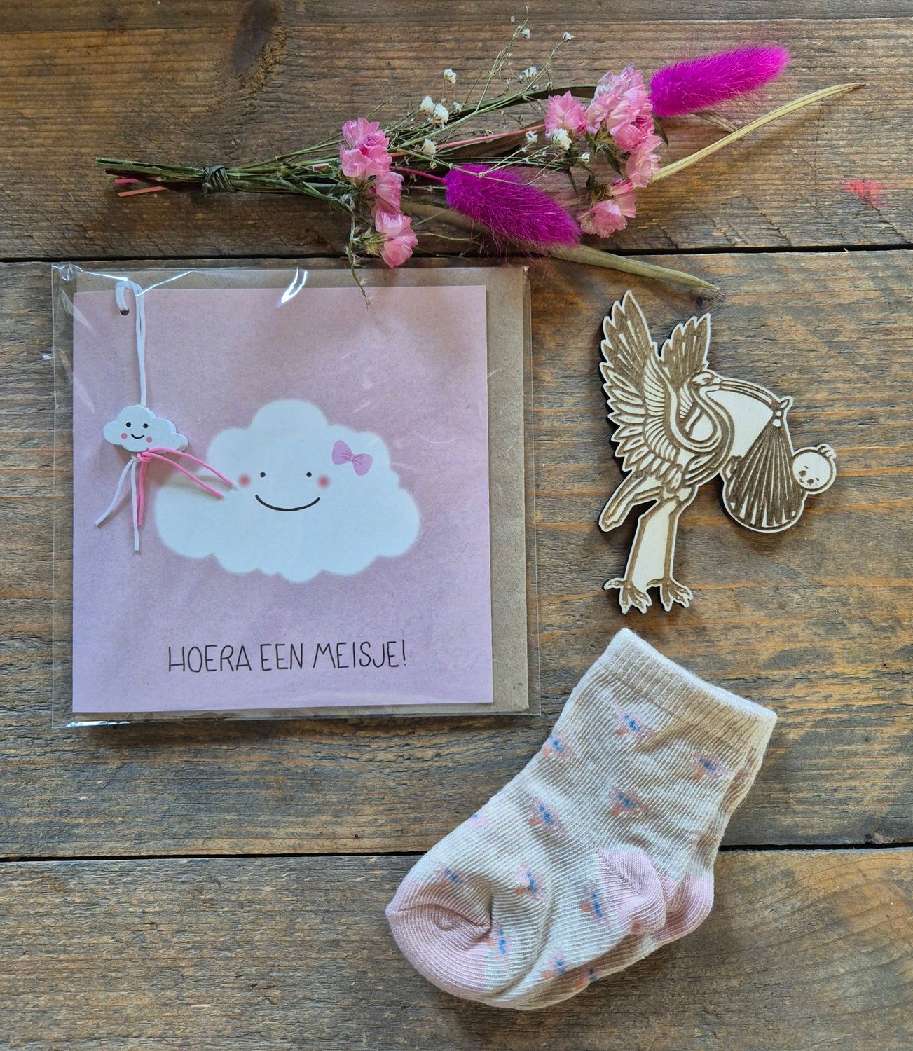 Plat gelegde afbeelding van een handgemaakte kaart, gedroogde bloemen, een gouden engelbroche en een pastelkleurige sok op een houten ondergrond.
