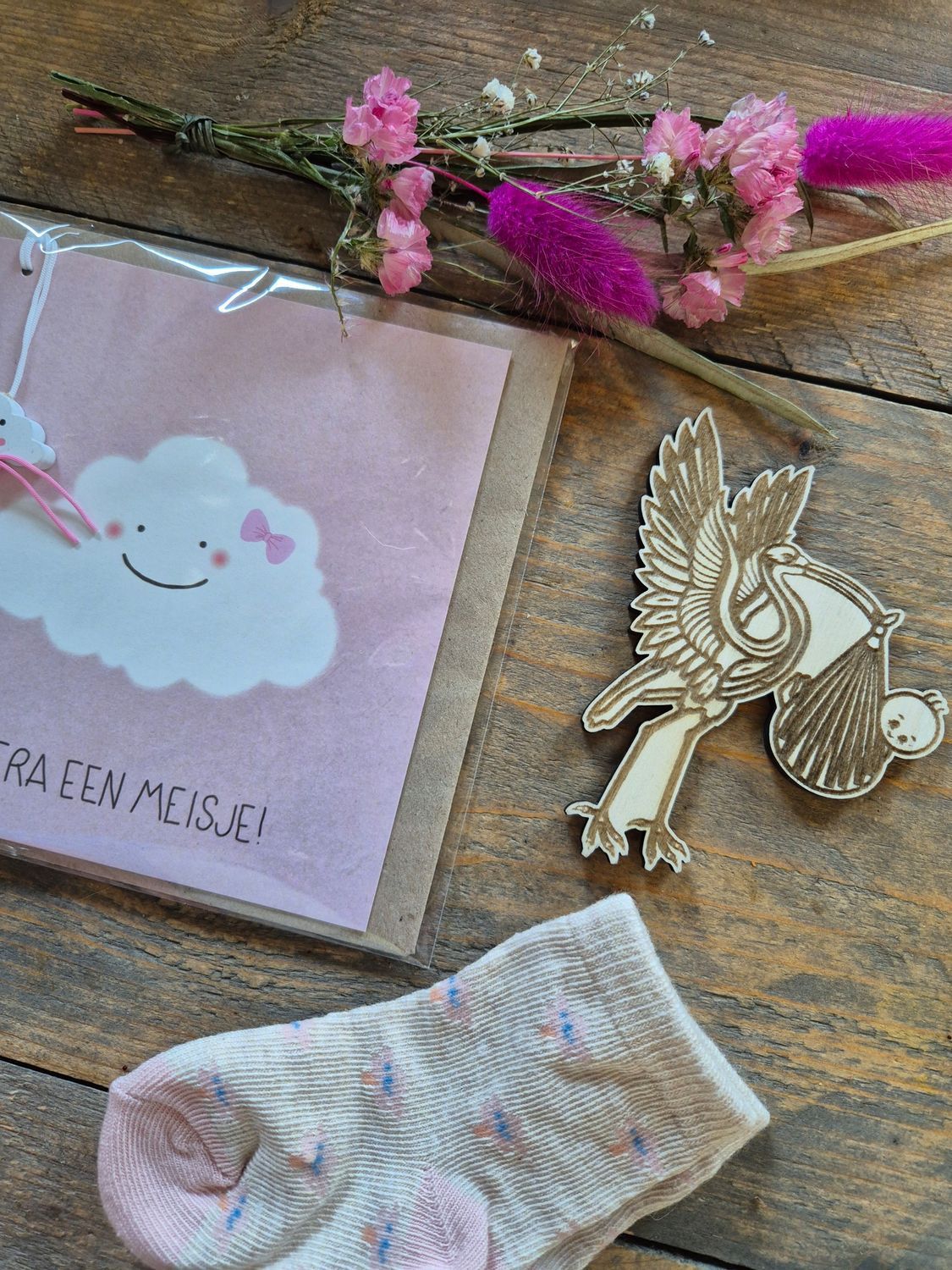 Plat gelegde afbeelding van een handgemaakte kaart, gedroogde bloemen, een gouden engelbroche en een pastelkleurige sok op een houten ondergrond.
