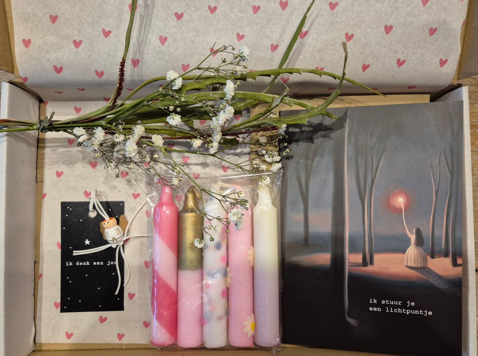 Cadeauverpakking met roze kaarsen, gedroogde bloemen en twee geïllustreerde kaarten met een zonsondergang en een sterrenhemel.