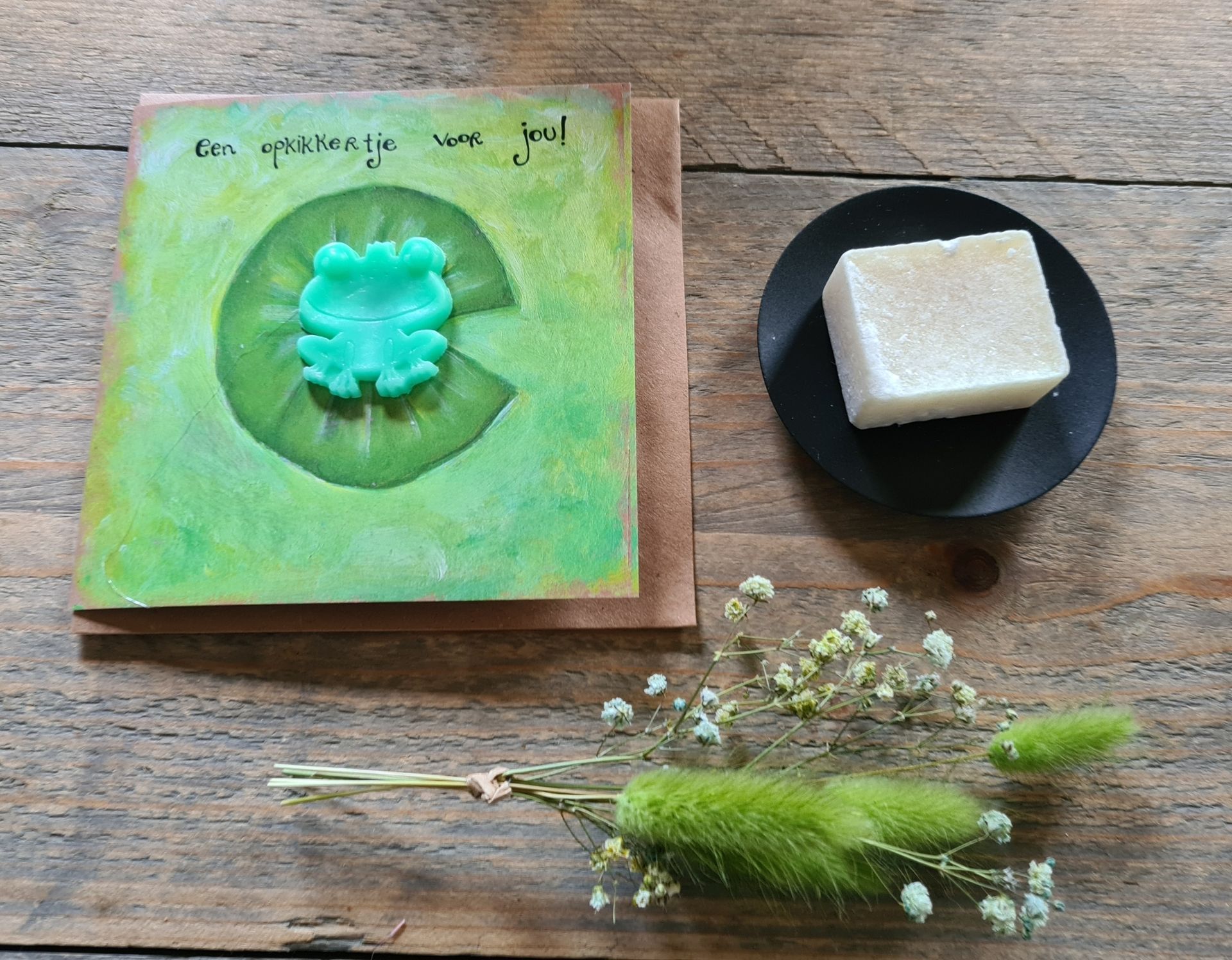Een groen stuk zeep met bloemenmotief op een zwart schaaltje, met een bijpassend zeepdoosje en gedroogde bloemen op een houten tafel.
