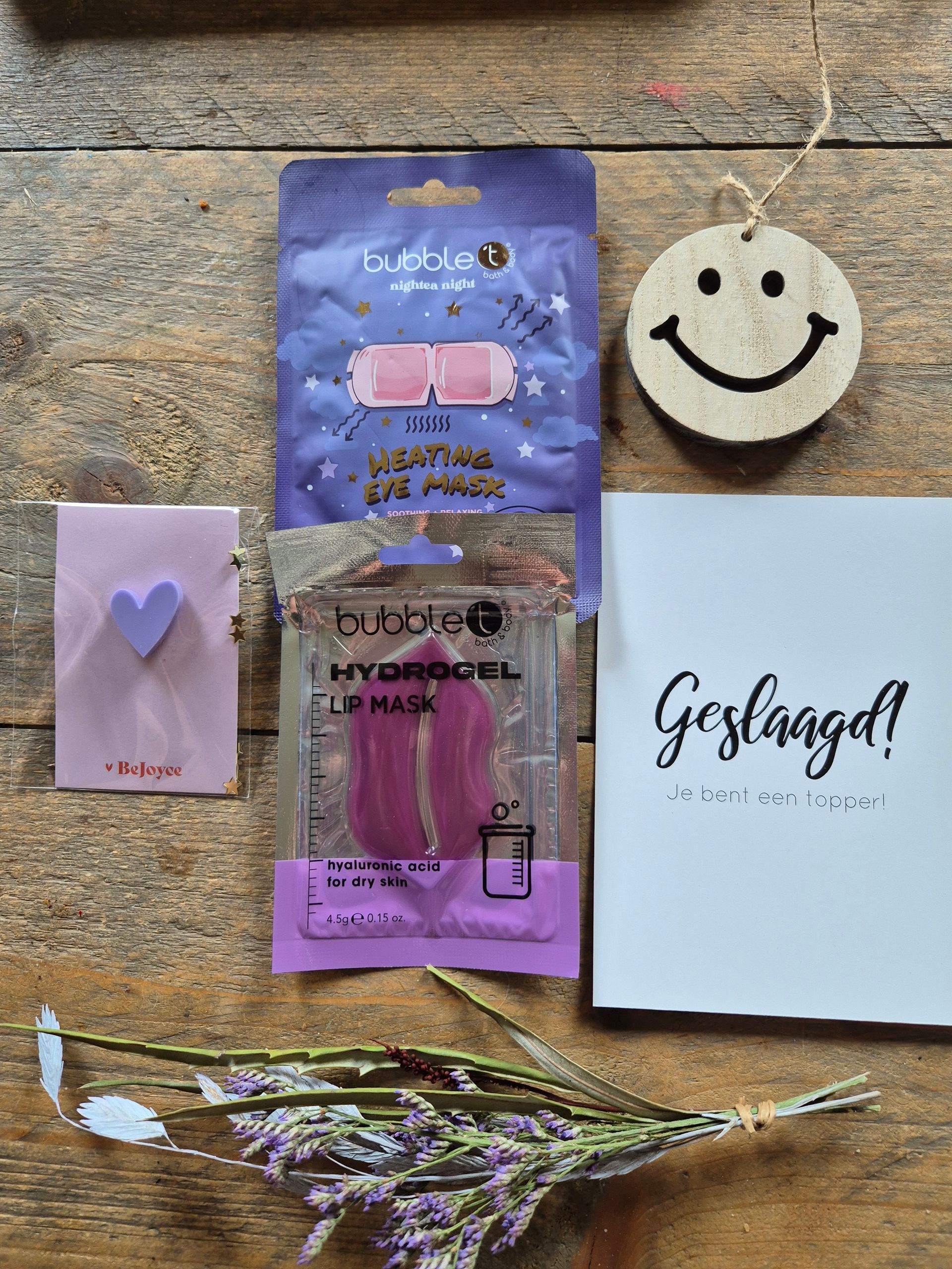 Diverse cosmetica- en cadeauartikelen op een bureau, waaronder een label met een smiley, een paars etui en gedroogde bloemen.