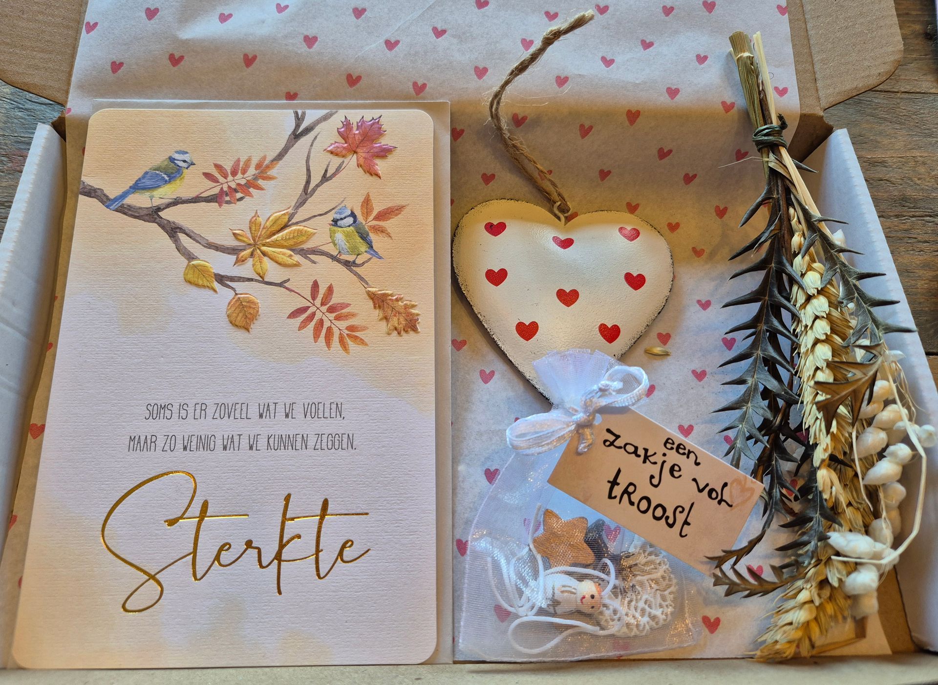 Cadeauverpakking met een notitieboekje met bloemenmotief, een hartvormige kaars, een snackzakje en gouden gedroogde bloemen, vastgebonden met een rood lint.