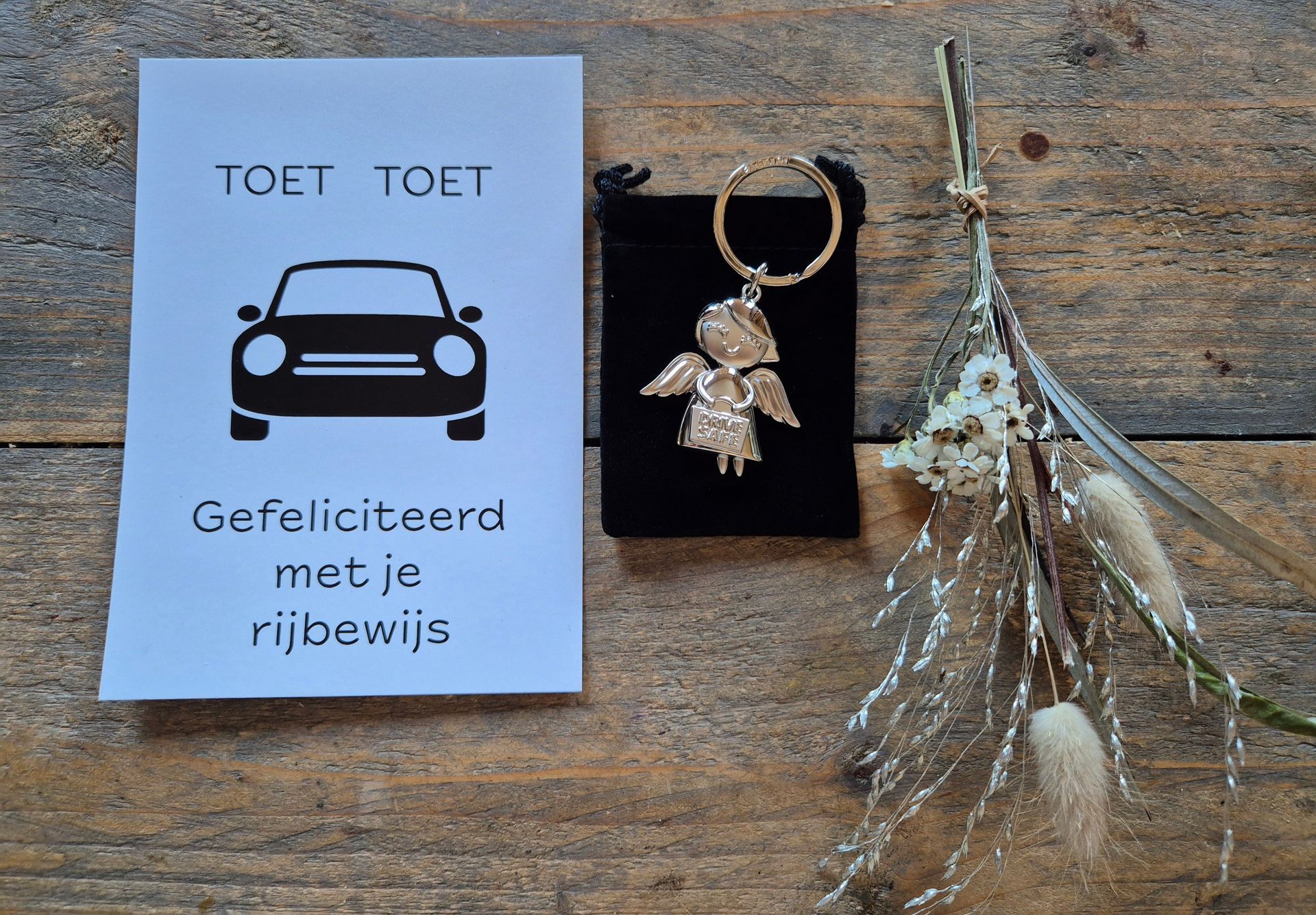 Cadeaukaart met auto-icoon en Nederlandse tekst naast een gouden engelsleutelhanger op een zwarte kaart, naast gedroogde bloemen.