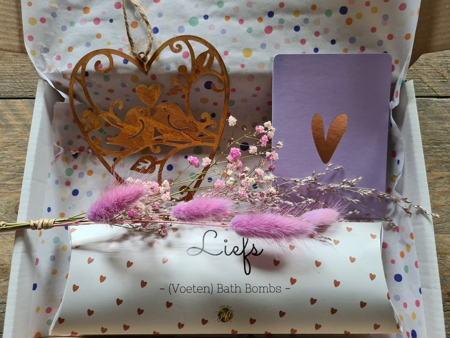Cadeauverpakking met pastelkleurige bloemen, paarse kaart en gouden hartjesversiering op een achtergrond met stippen.