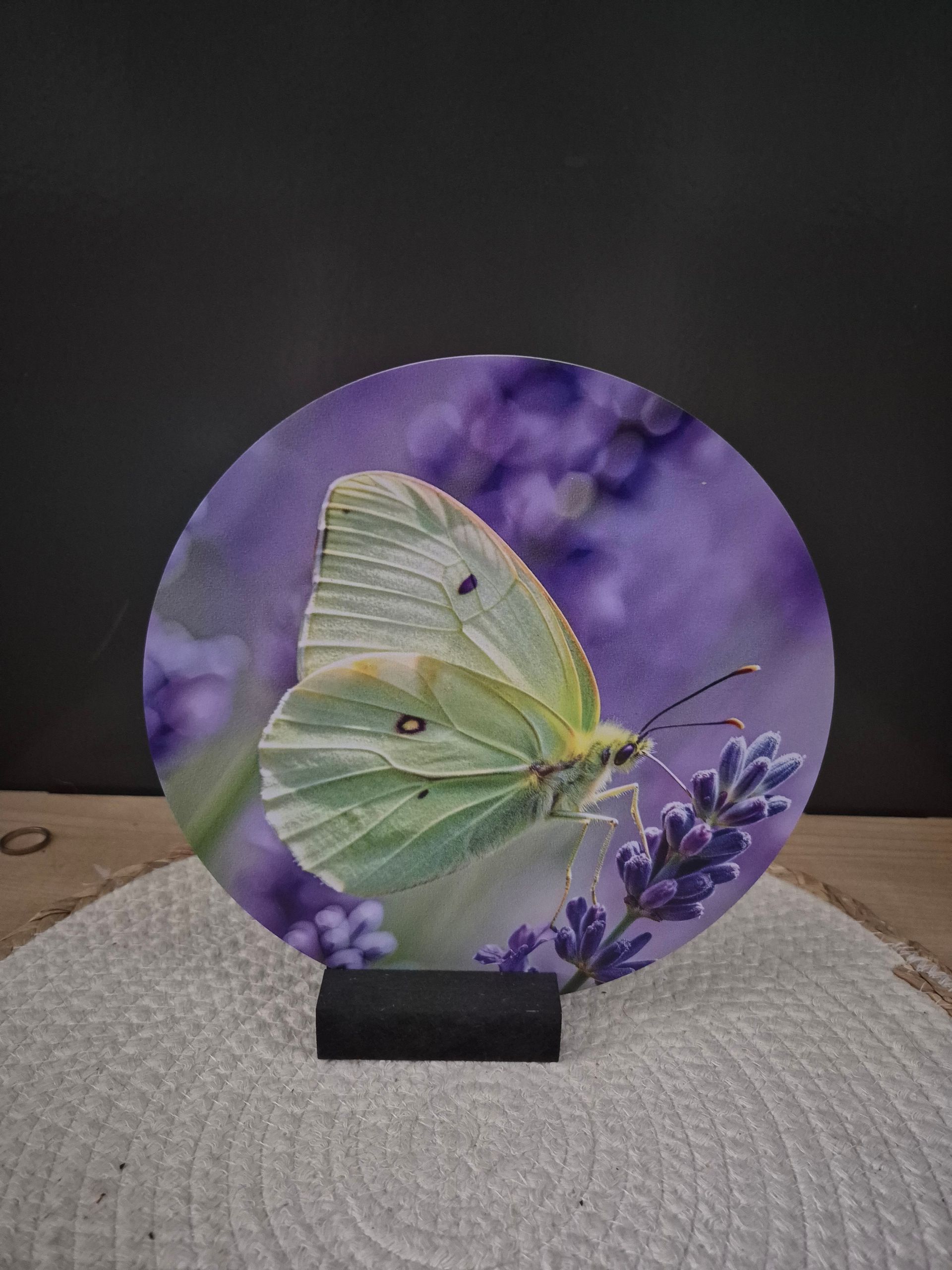 Een rond plaatje met een paarse vlinder erop, geplaatst op een klein zwart standaardje boven een geweven placemat.