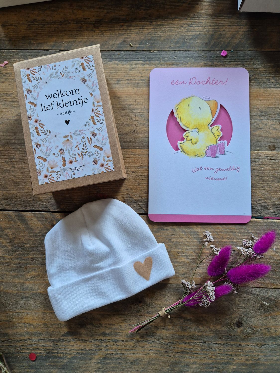 Een wit babymutsje, twee wenskaarten en paarse bloemen, gerangschikt op een houten ondergrond.