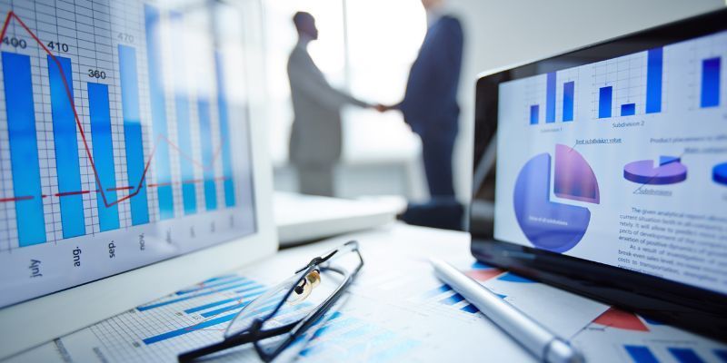 Riunione di lavoro con grafici su computer portatili e documenti su una scrivania, con persone sfocate che si stringono la mano sullo sfondo.