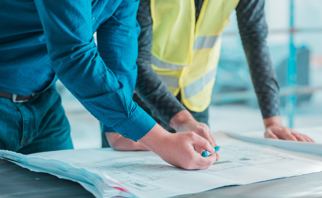 Due persone in abiti da lavoro esaminano i progetti su un tavolo in un cantiere.